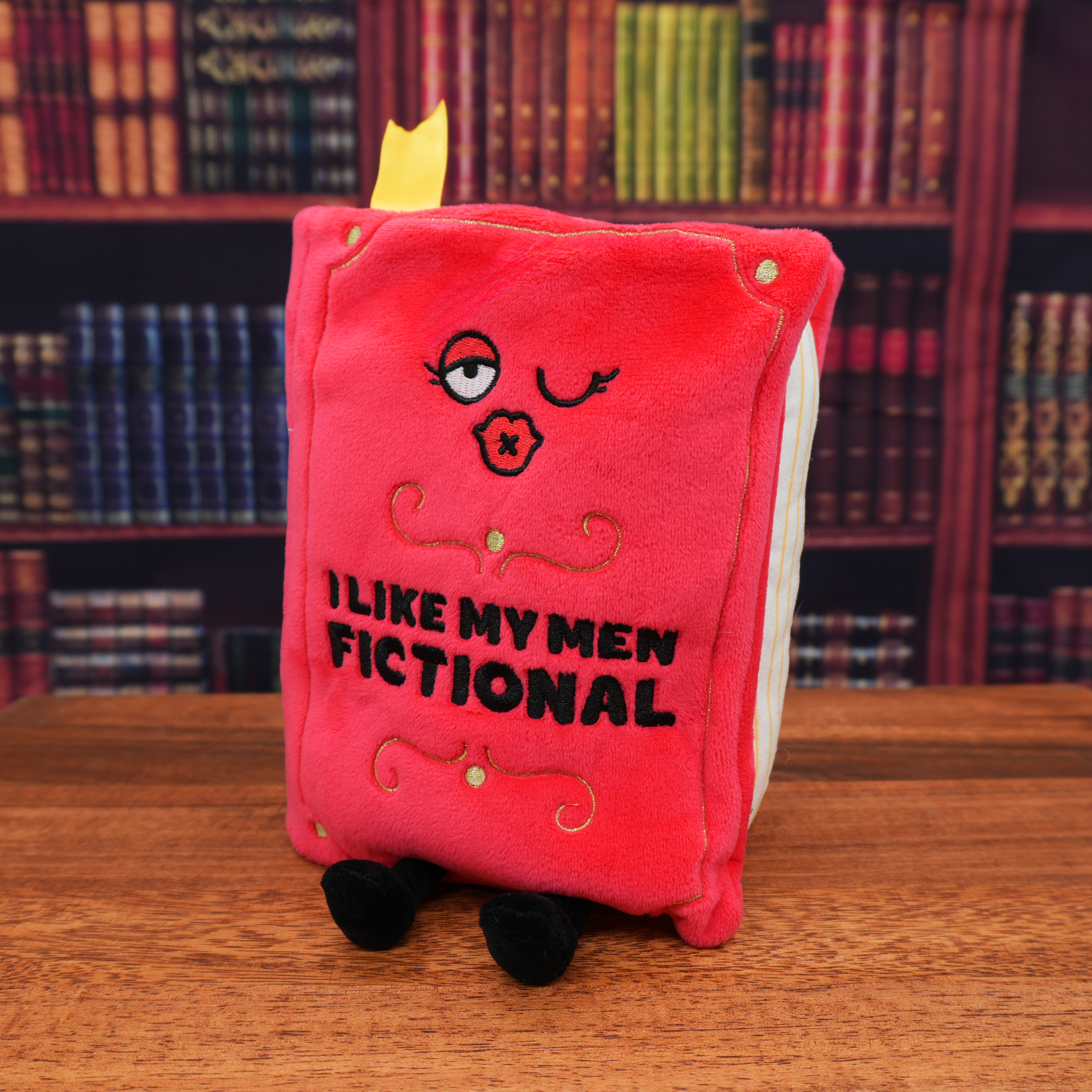 A red plush book with 'I LIKE MY MEN FICTIONAL' printed on the cover. The plush has a face and a pair of legs and is placed on a wooden surface, bookshelf background.