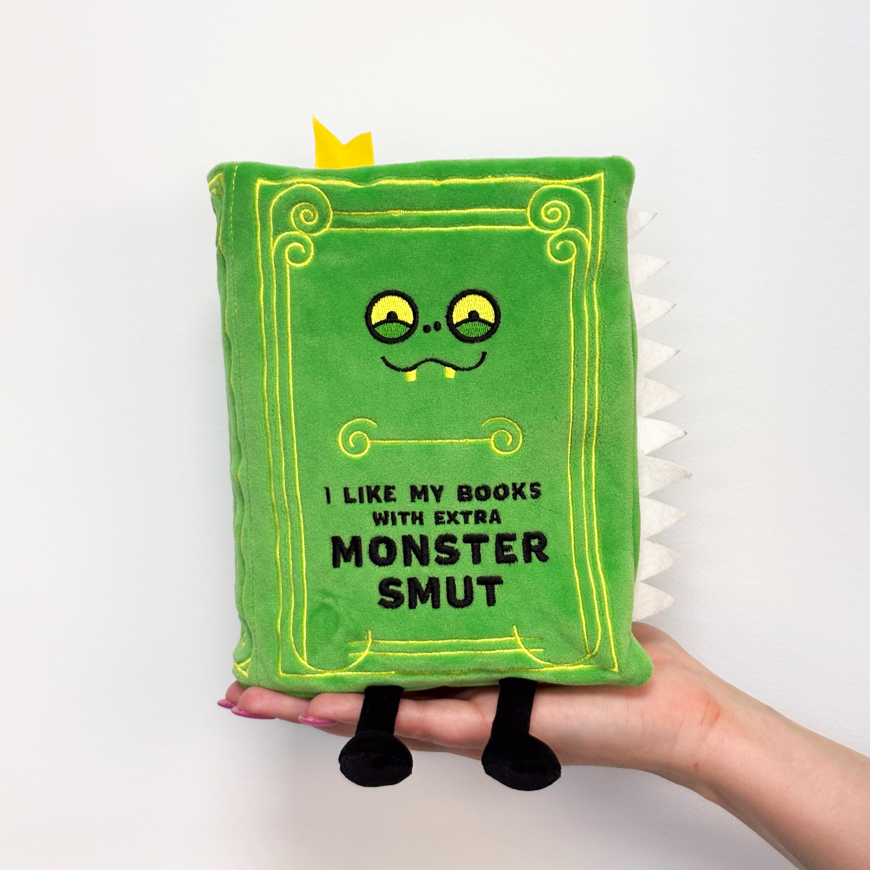 Hand holding a book plush with 'I like my books with extra monster smut' embroidered on the cover.