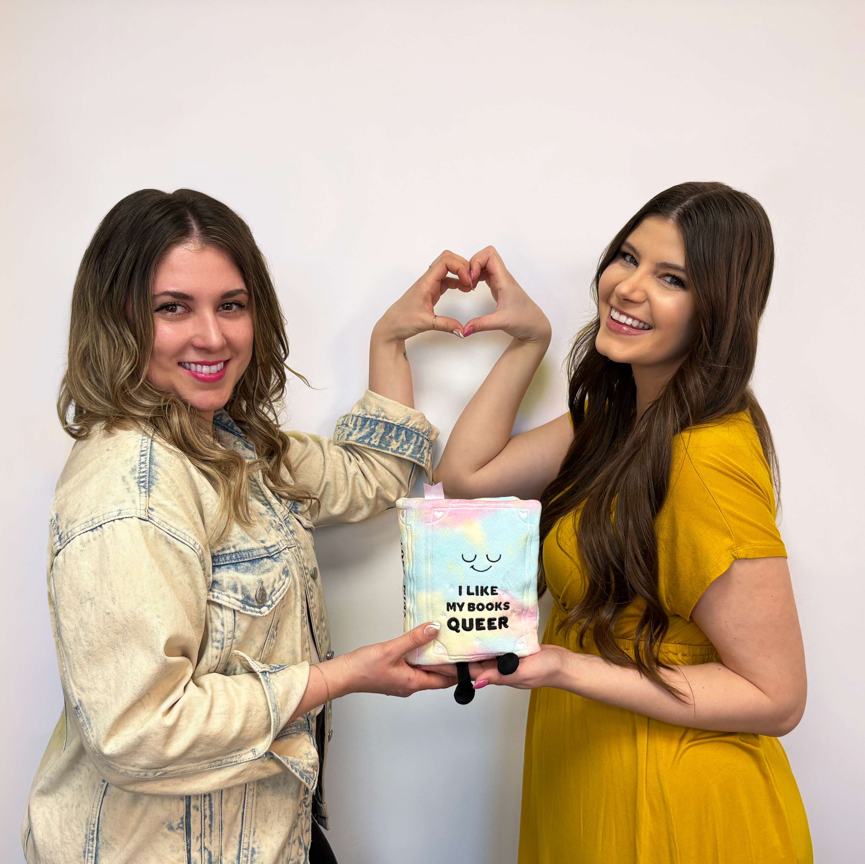 Two girls making a heart shape with their hands while holding a book that reads 'I like my book queer' in the other.