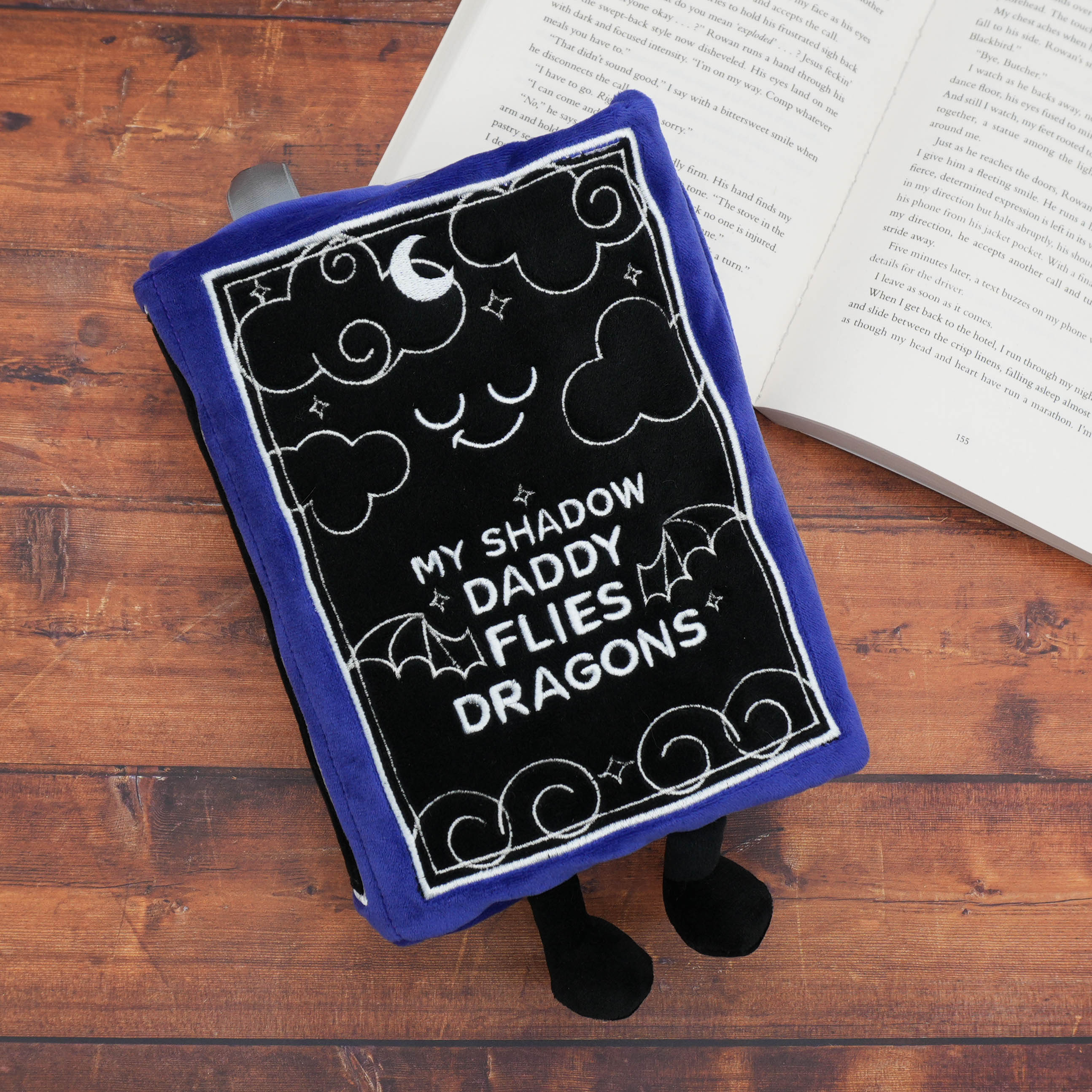 Plushie shaped like a book with 'my shadow daddy flies dragons' text on a wooden surface