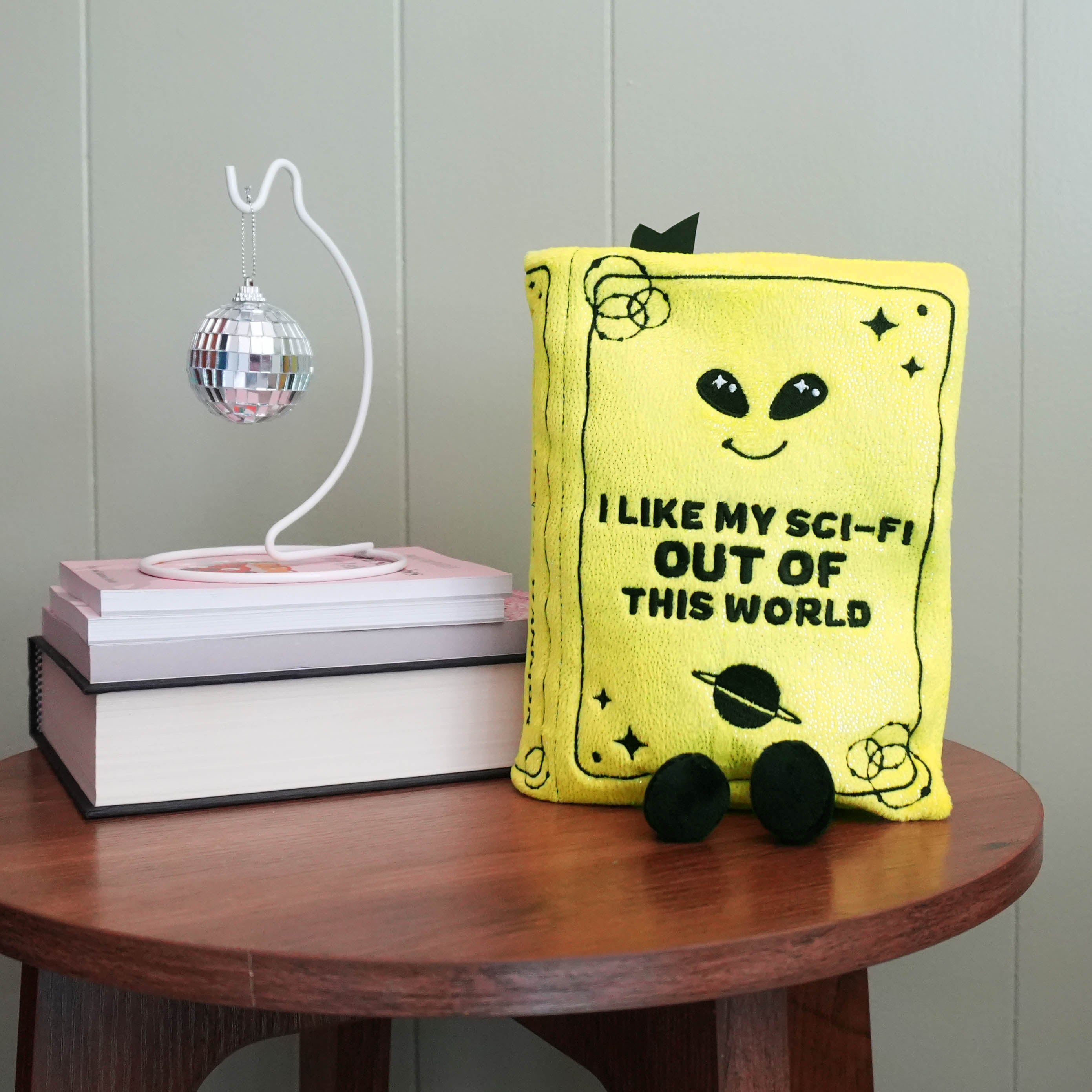Yellow pillow with alien design and text on a wooden table with books and a disco ball.