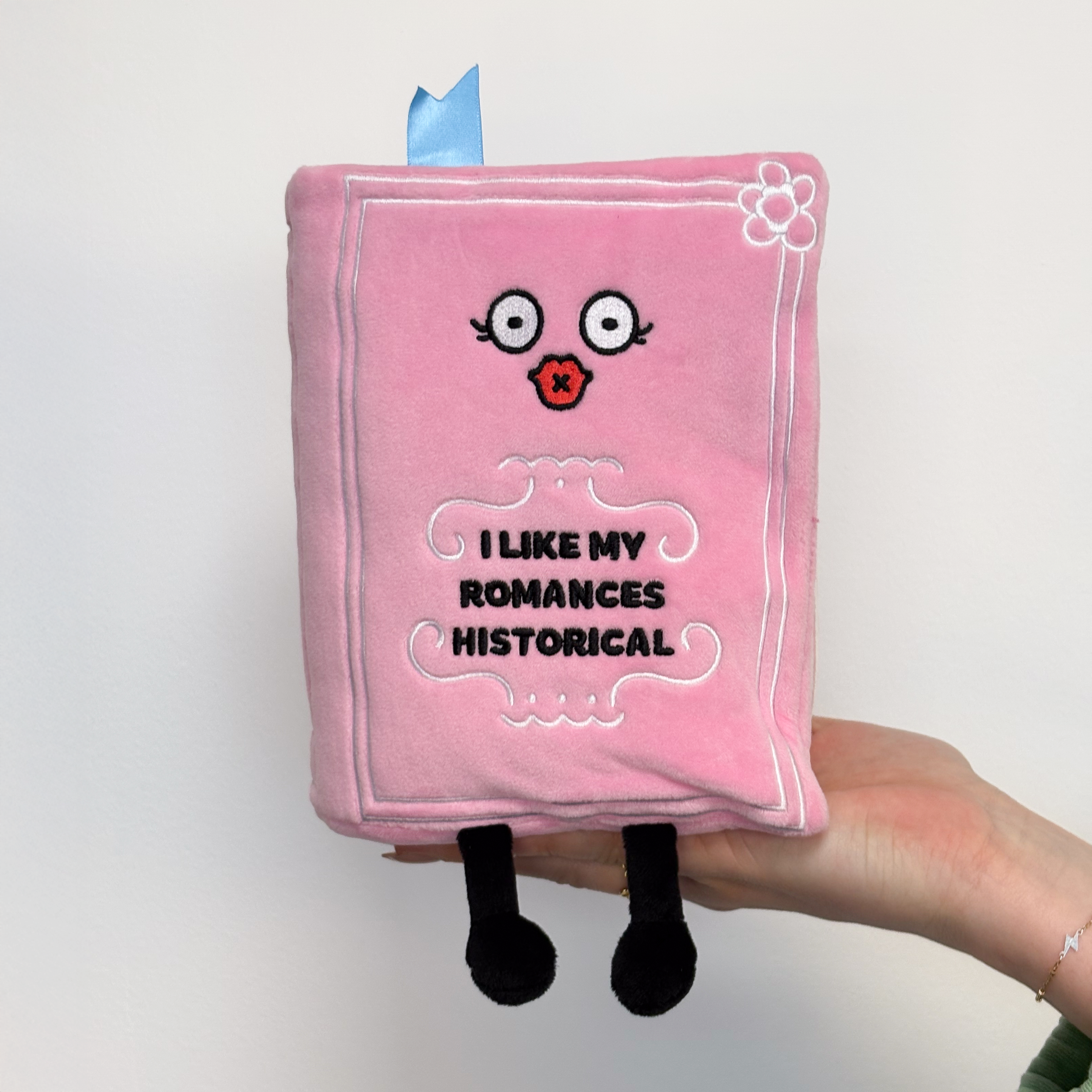 A hand holding a pink plush designed to look like a book with a face, dangly legs, and text that reads, 'I like my romances historical'.