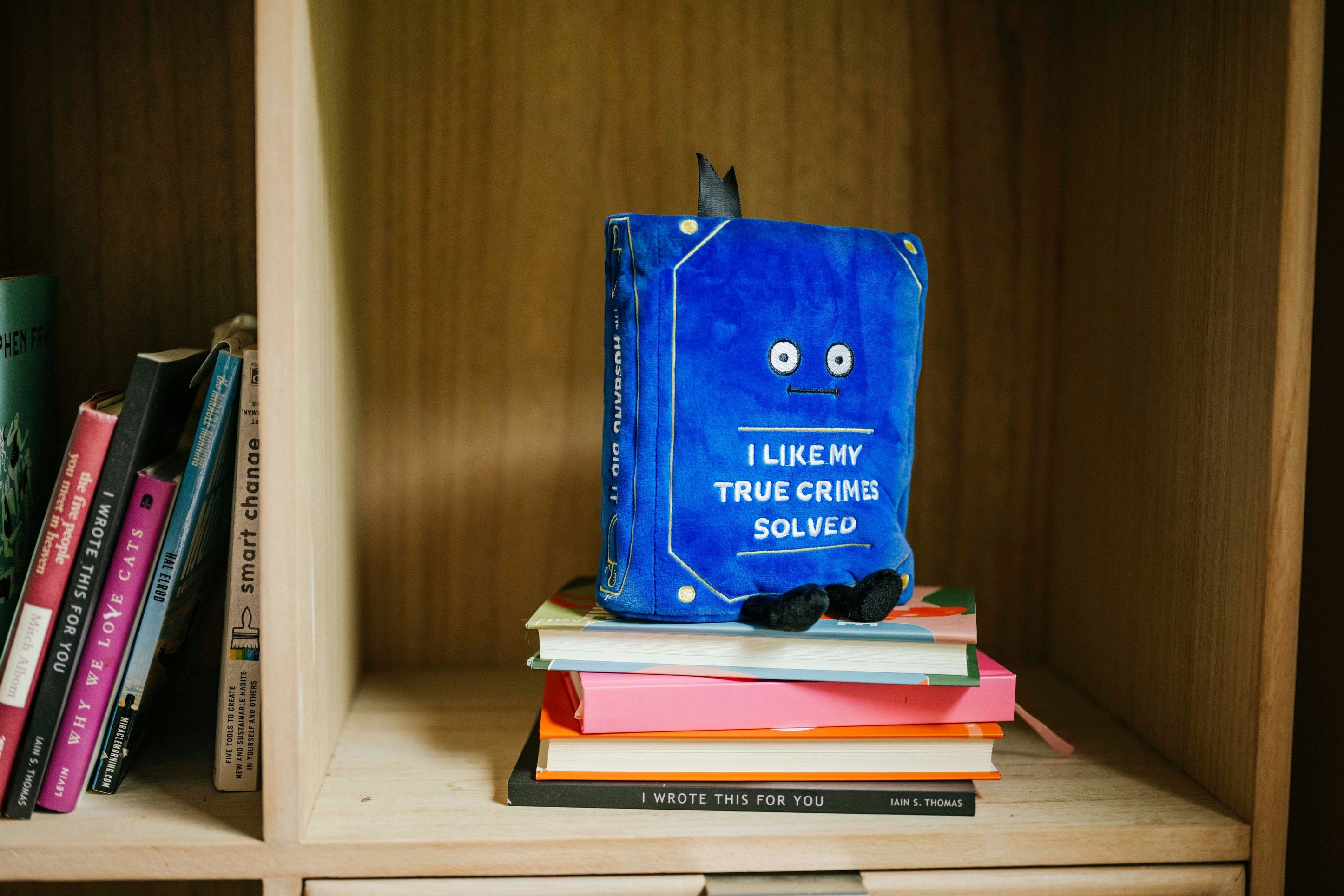 A plush designed to look like a book with the title 'I LIKE MY TRUE CRIMES SOLVED' on the cover placed on a bookshelf.