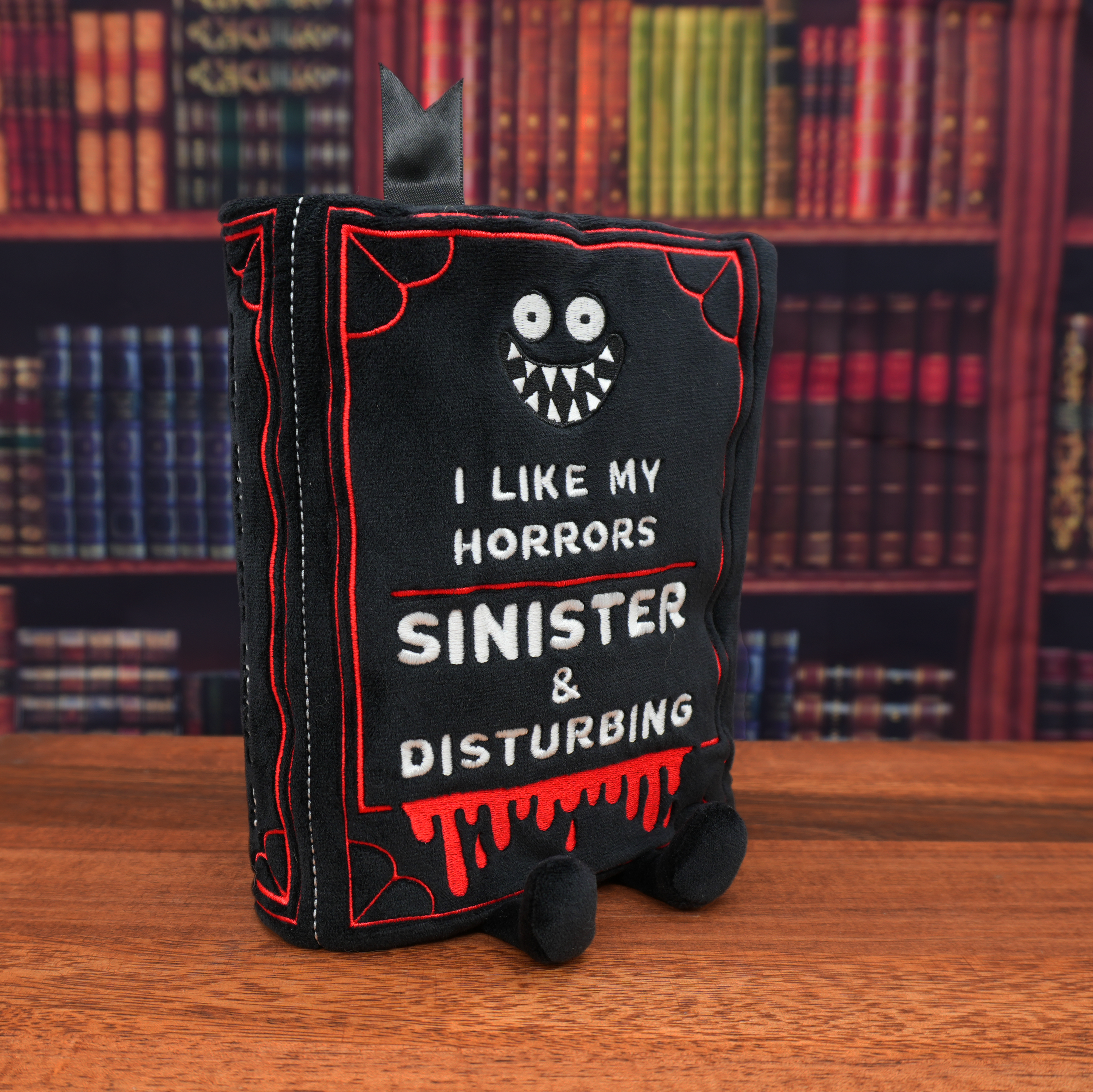 A plush book featuring the text 'I LIKE MY HORRORS SINISTER & DISTURBING' on the front in front of a library wall.