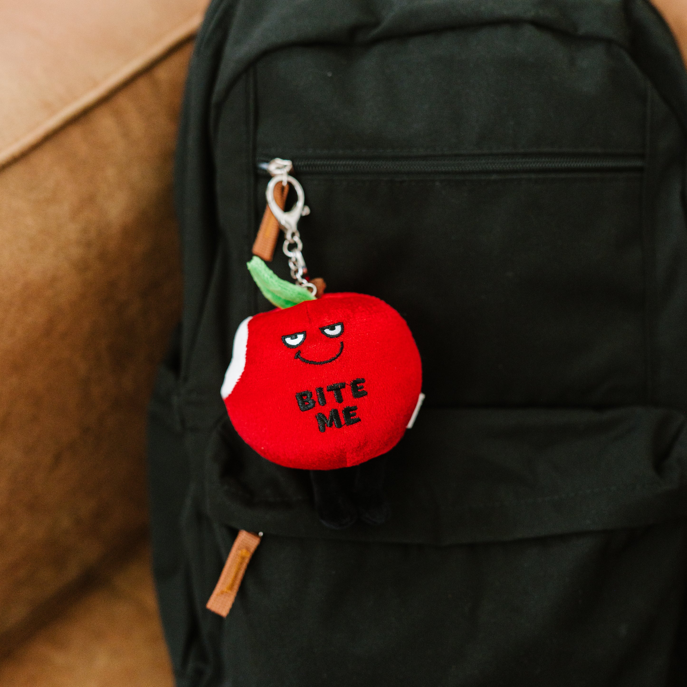 A plush bag charm designed to look like a red apple with a happy face and text saying 'BITE ME' hanging off a backpack.