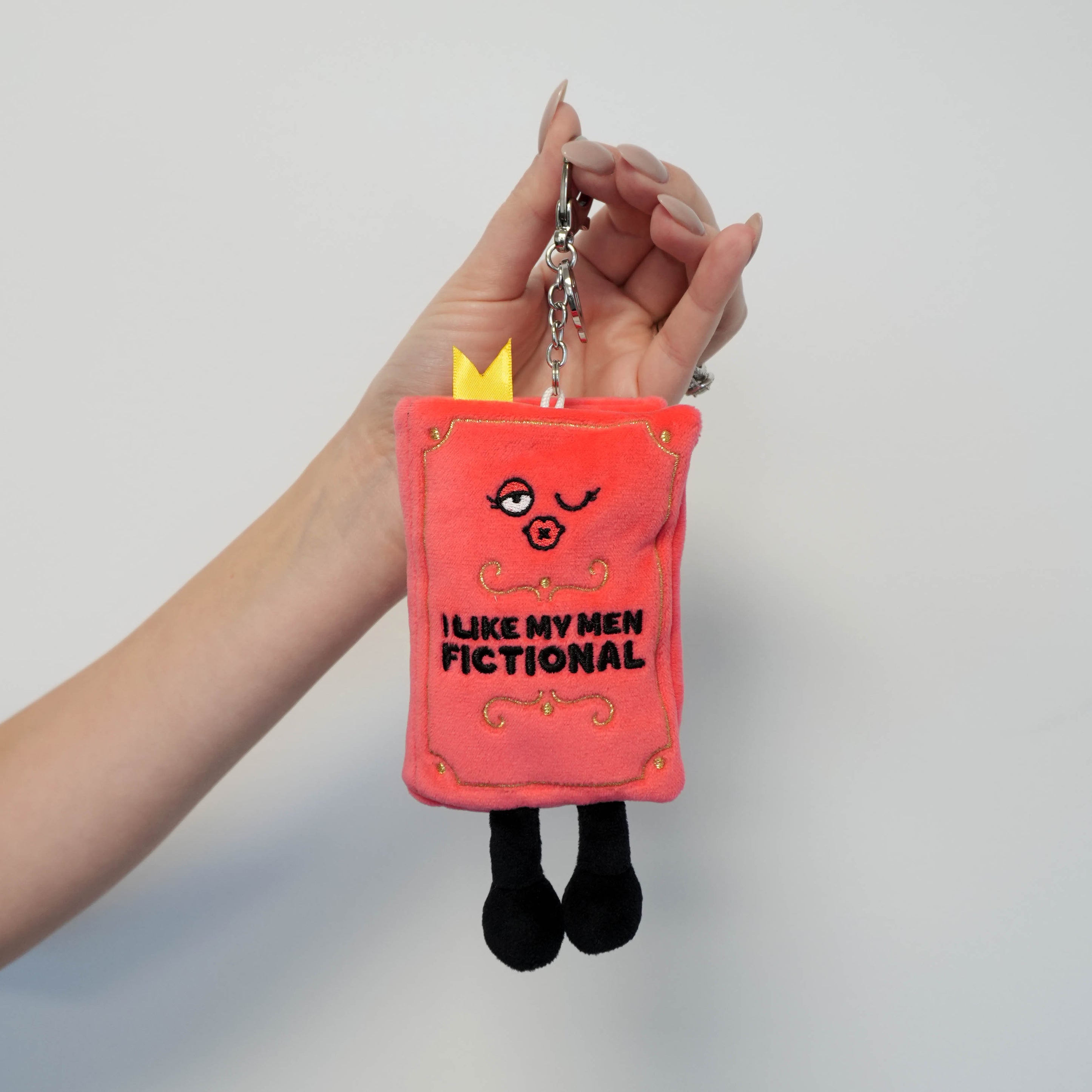 Plush bag charm with cartoon face and 'I like my men fictional' text held by a hand against a gray background