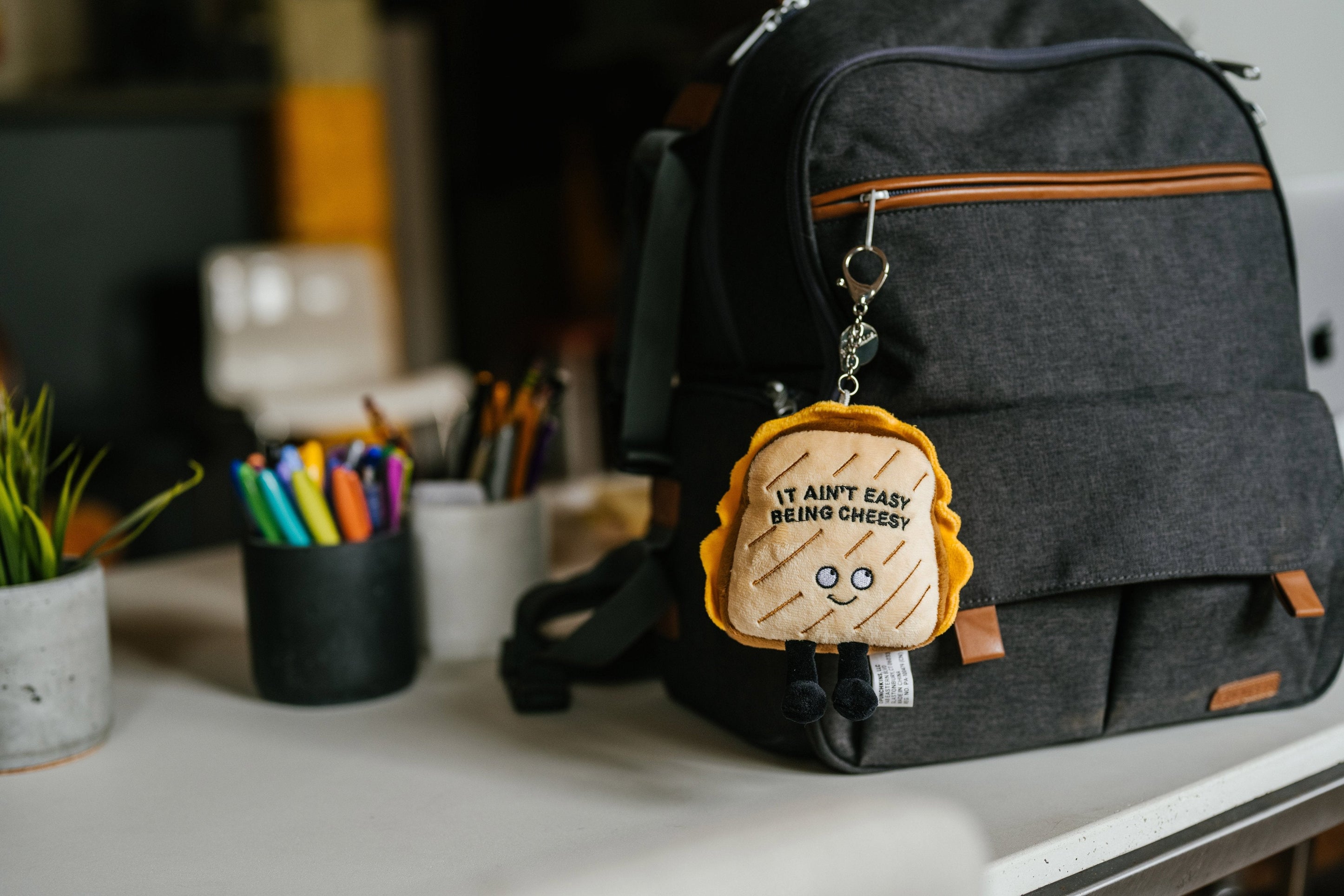 A plush charm designed to look like a grilled cheese sandwich with the phrase 'It Ain't Easy Being Cheesy' written on it hanging on a backpack.