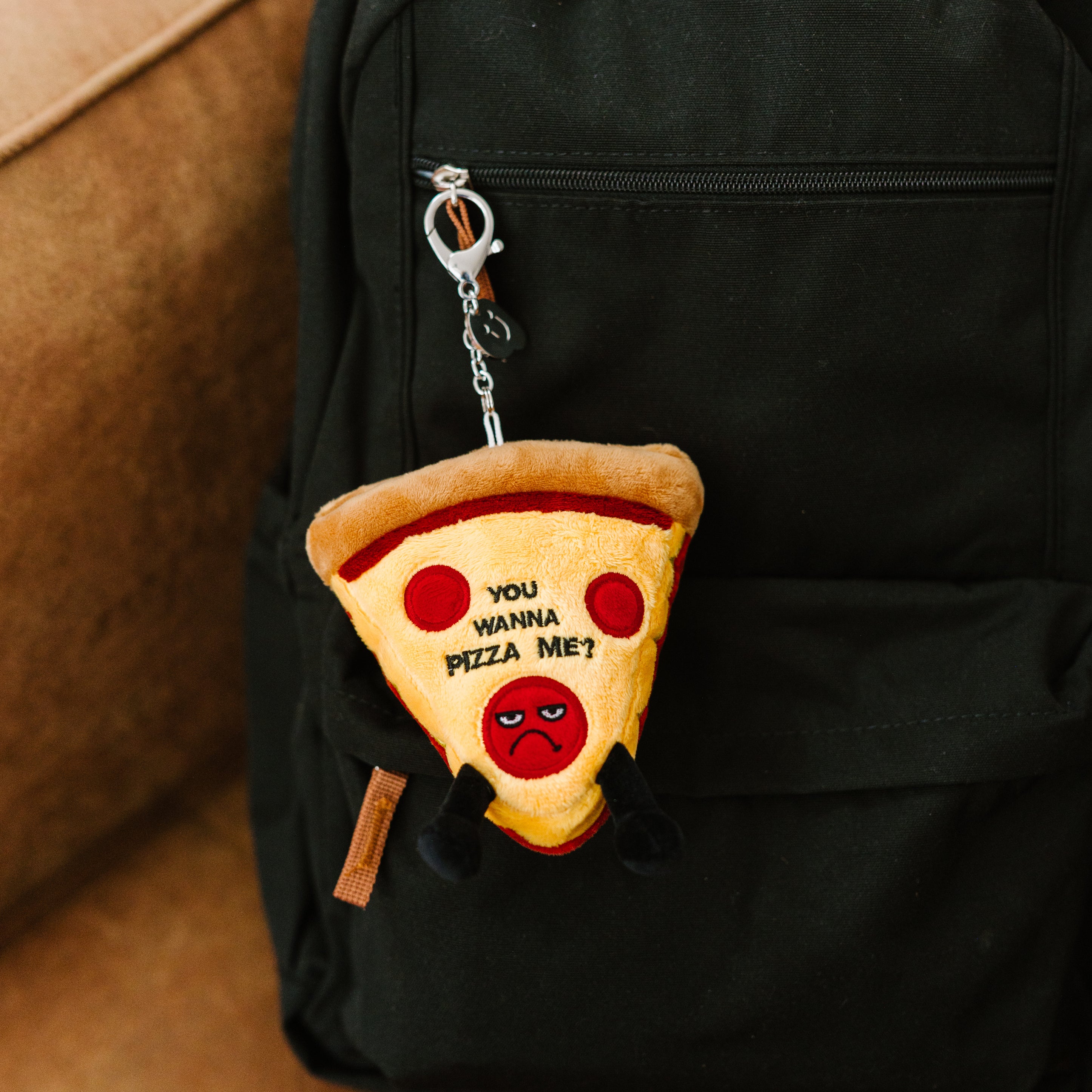 A plush shaped like a slice of pizza with an angry expression, text 'YOU WANNA PIZZA ME?', and dangling legs. It's hanging off a backpack.