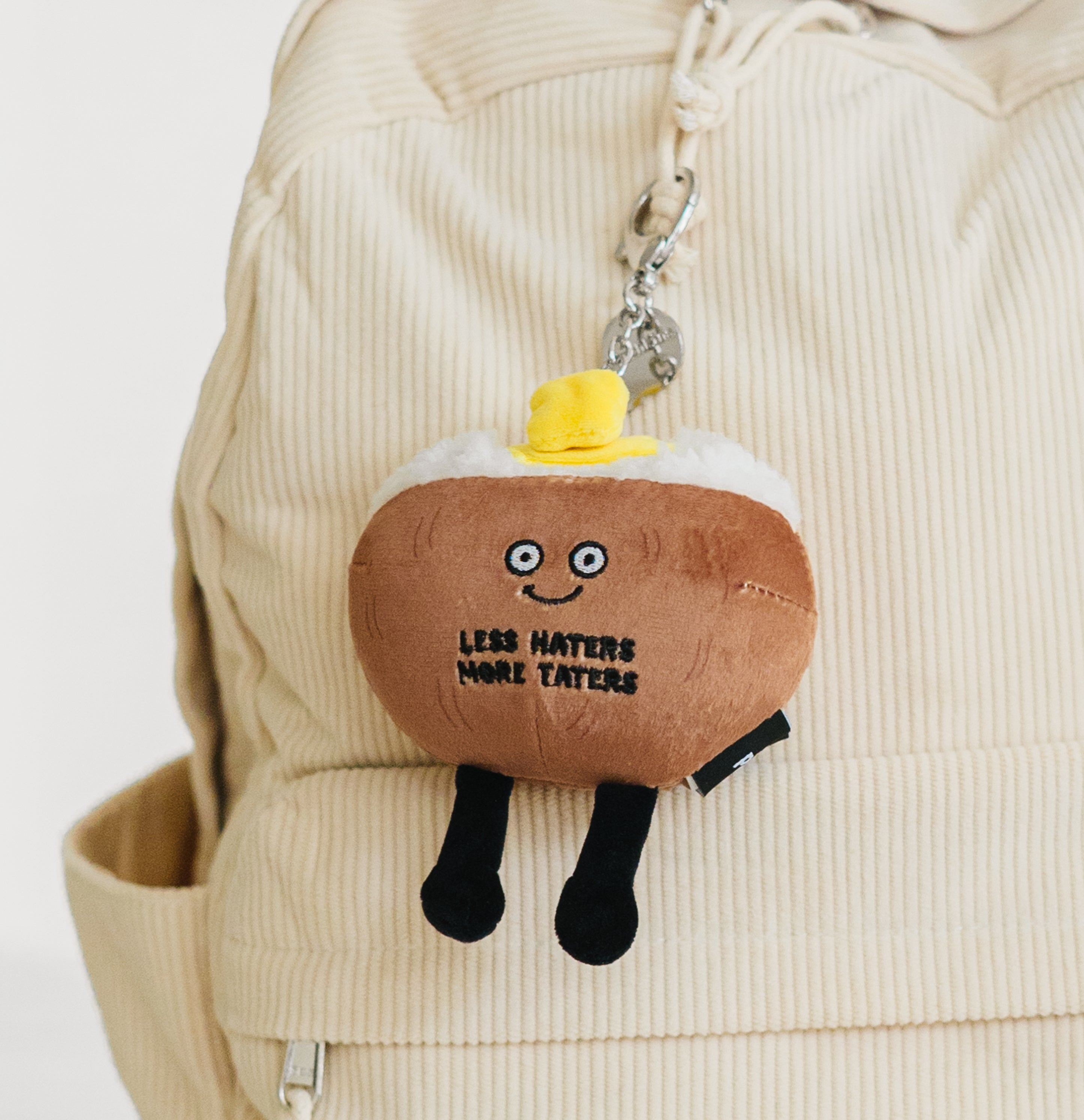 A plush bag charm designed to look like a baked potato with the text 'LESS HATERS MORE TATERS' printed on it hanging from a backpack.