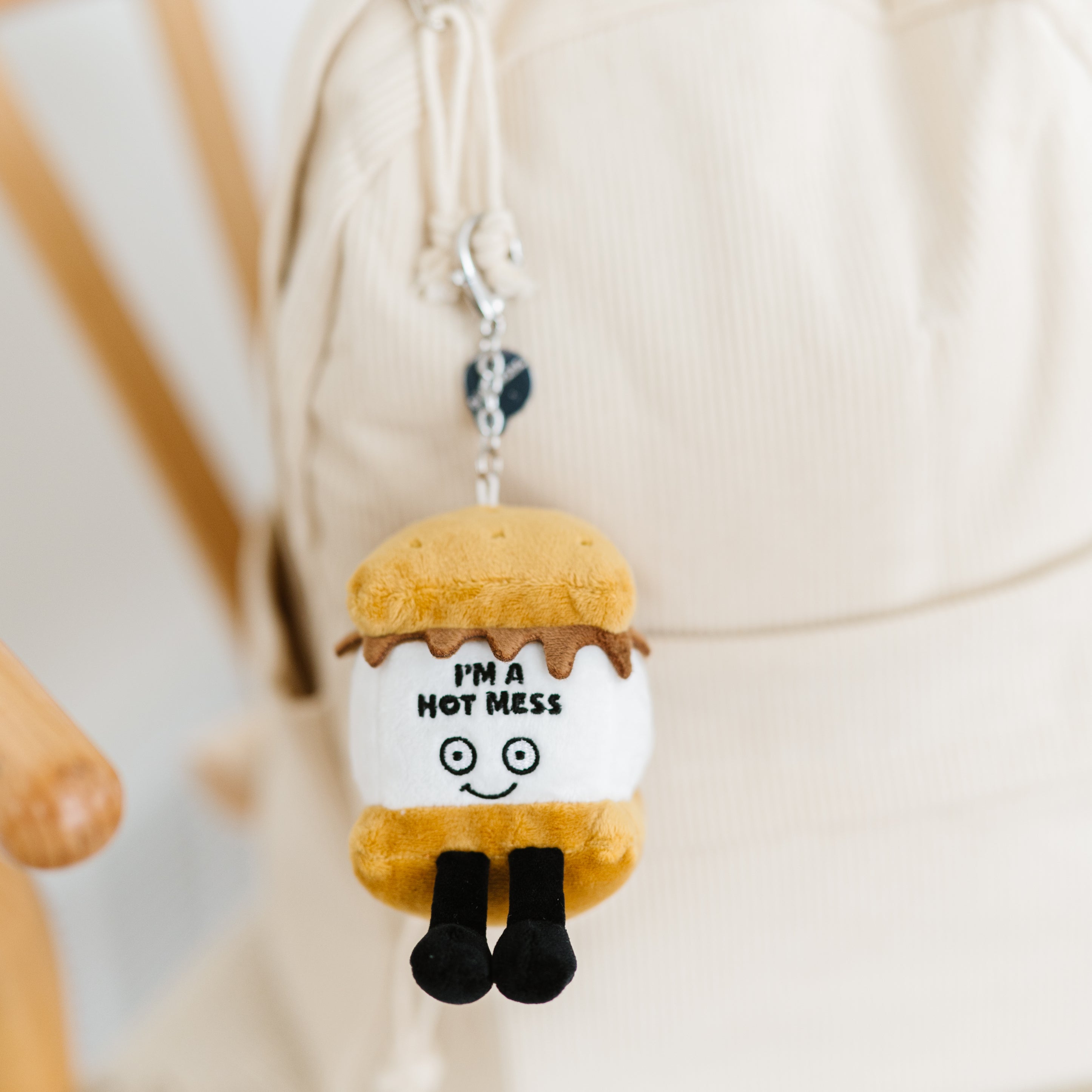 A plush bag charm designed to look like a s'more with the printed text 'I'M A HOT MESS' on it hanging on a backpack.