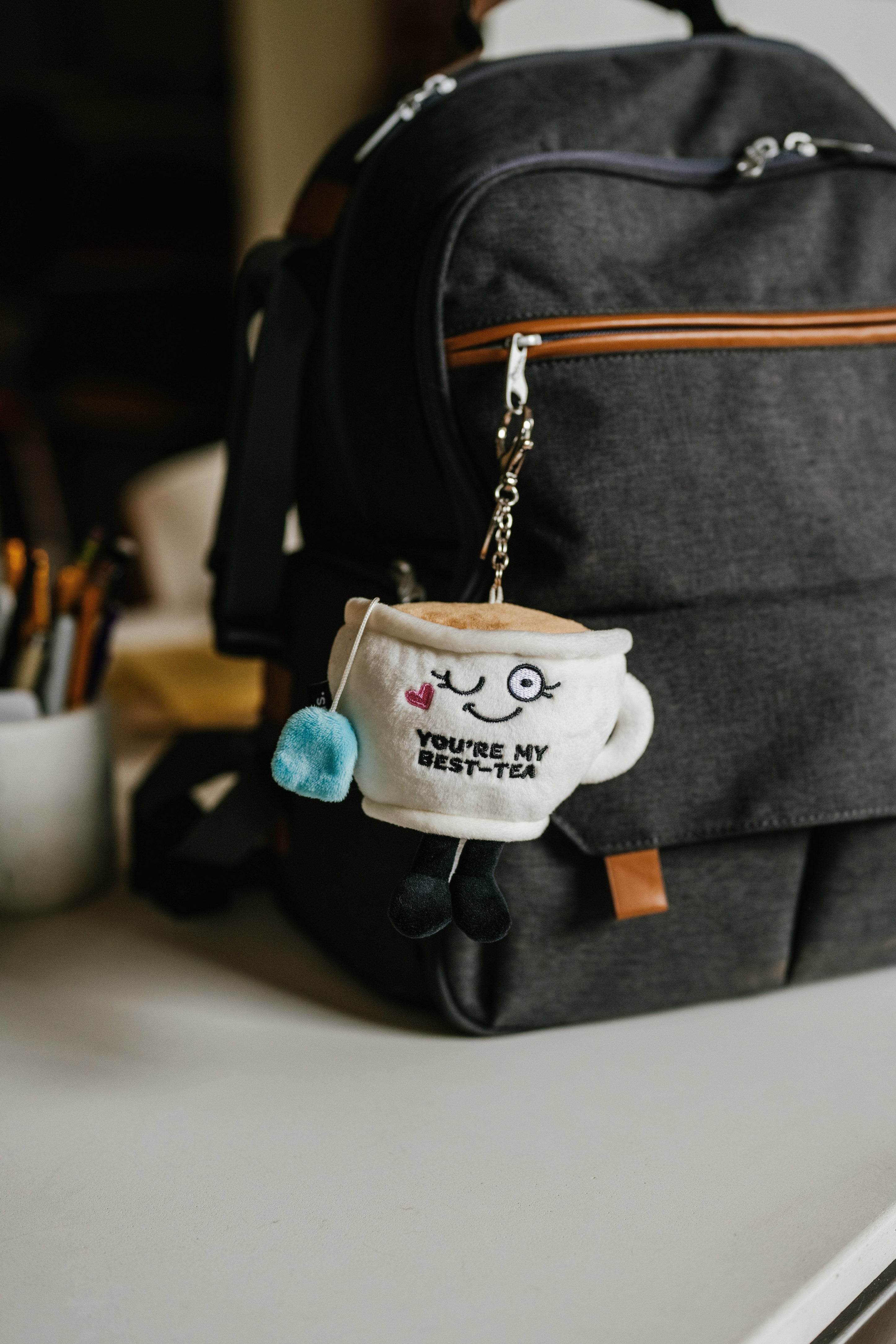 A plush teacup bag charm with a cute face and 'you're my best-tea' text embroidered on it hanging on a backpack.