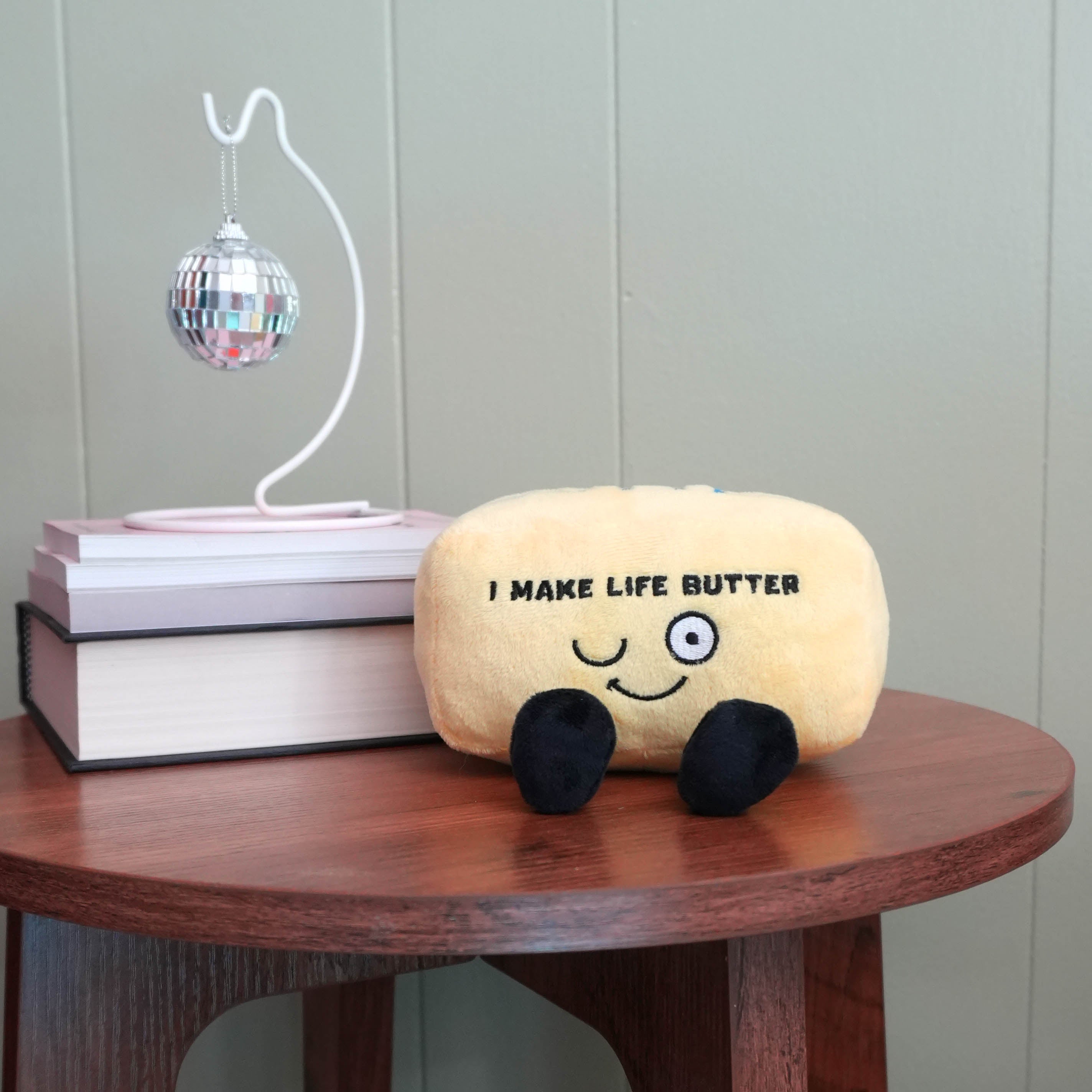 Plush toy resembling a stick of butter with text on a wooden table, books, and a disco ball in the background.