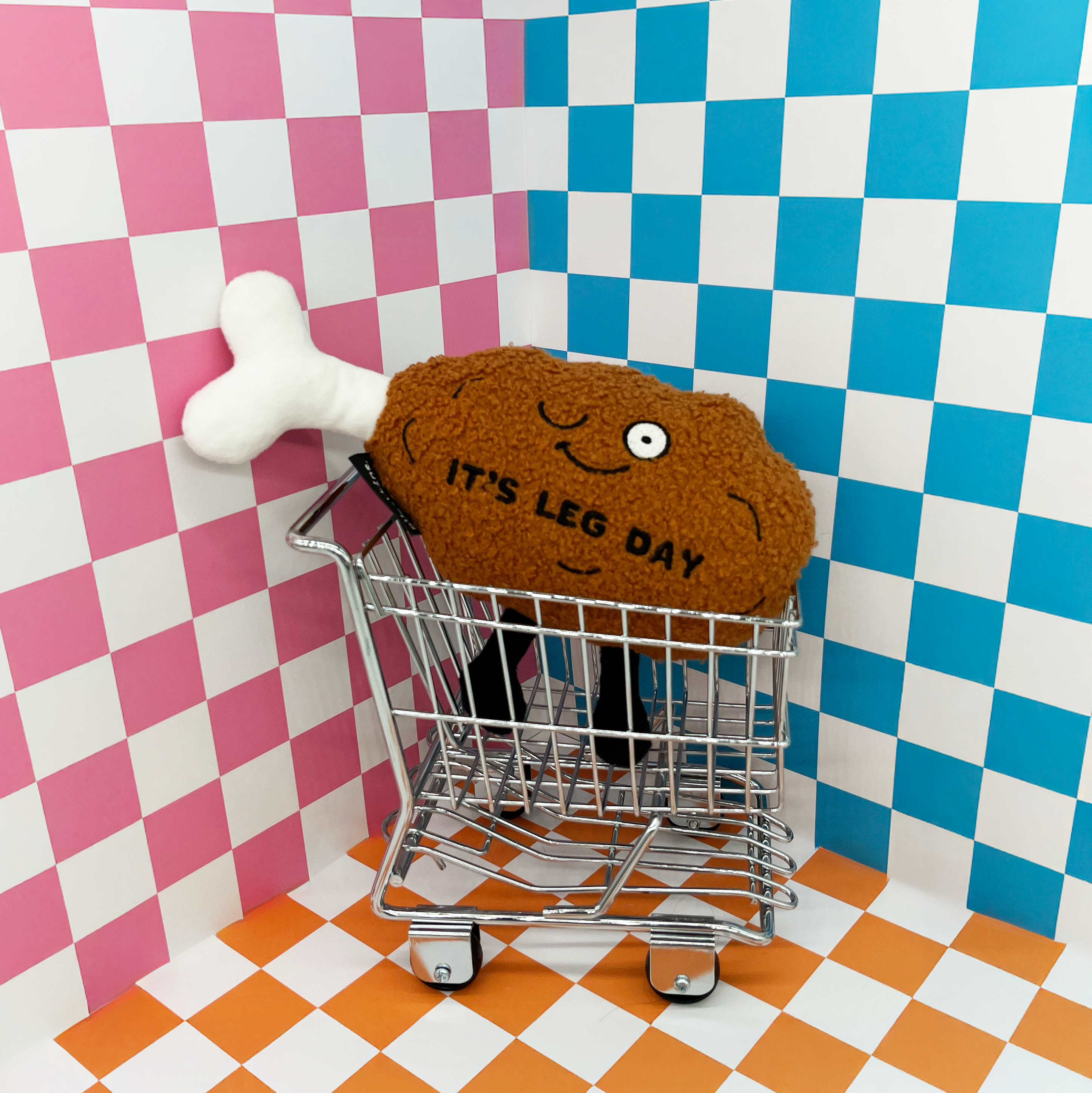 A plush chicken leg with a smiling face and the text 'IT'S LEG DAY' on it placed in a shopping cart on a checkered background.