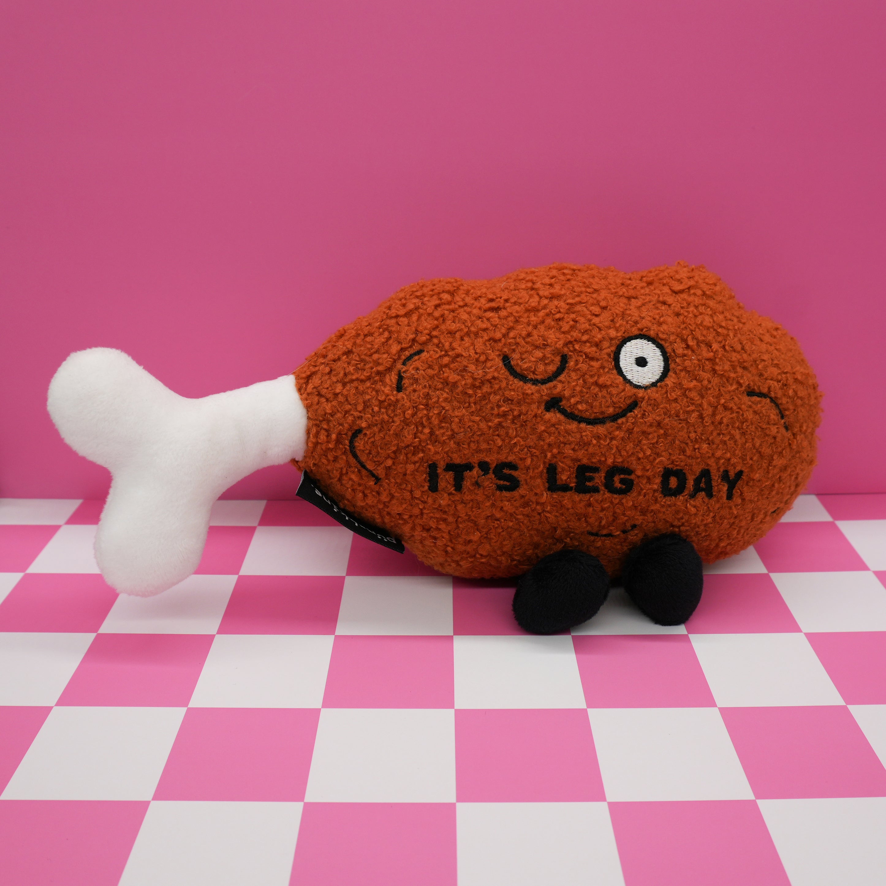 Plush toy resembling a chicken leg with a face and 'It's Leg Day' text on a pink and white checkered background.