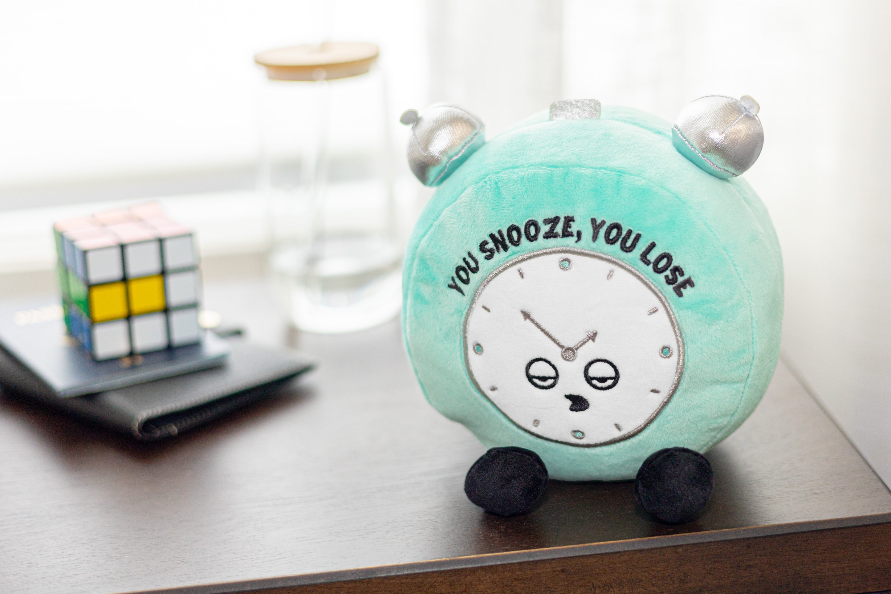 A plush alarm clock toy with the phrase 'YOU SNOOZE, YOU LOSE' written on the clock face. It's placed on a dresser.