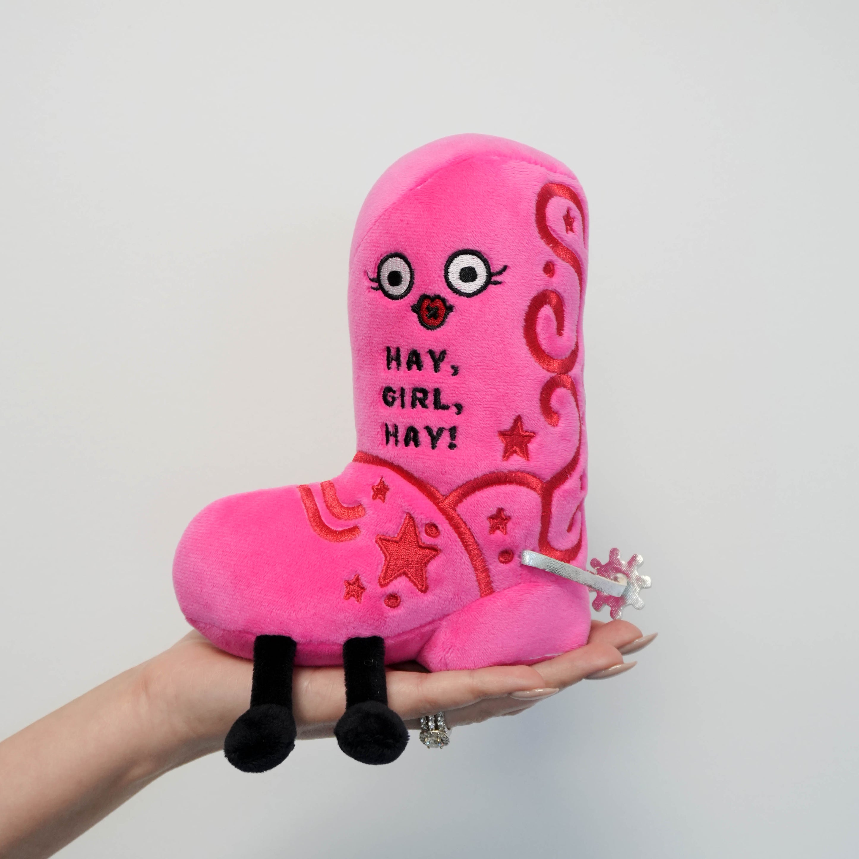 Pink plush toy with text 'Hay, Girl, Hay!' held in a hand against a white background.