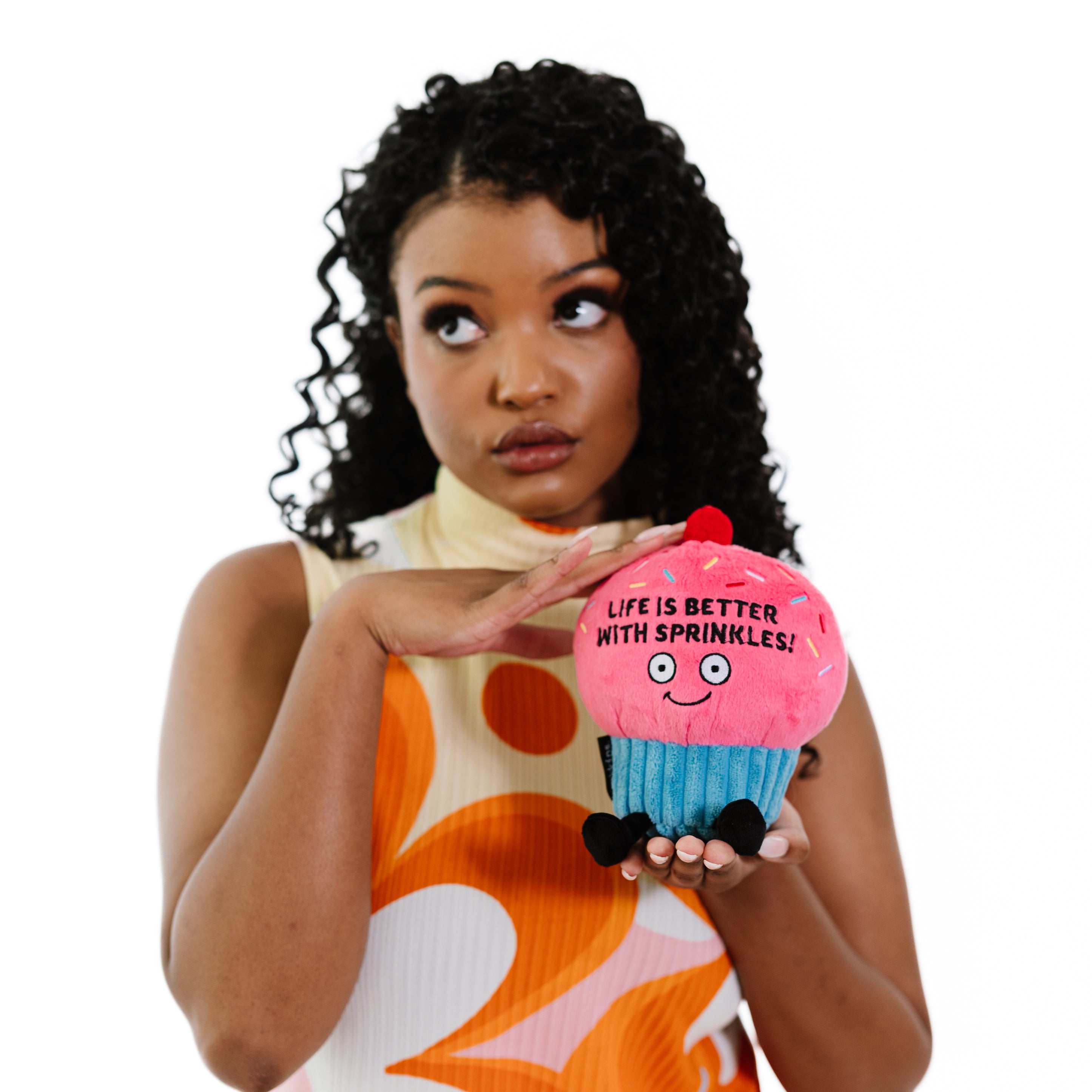 A woman holding a plush shaped like a cupcake with pink frosting with the phrase 'LIFE IS BETTER WITH SPRINKLES!' written on it.