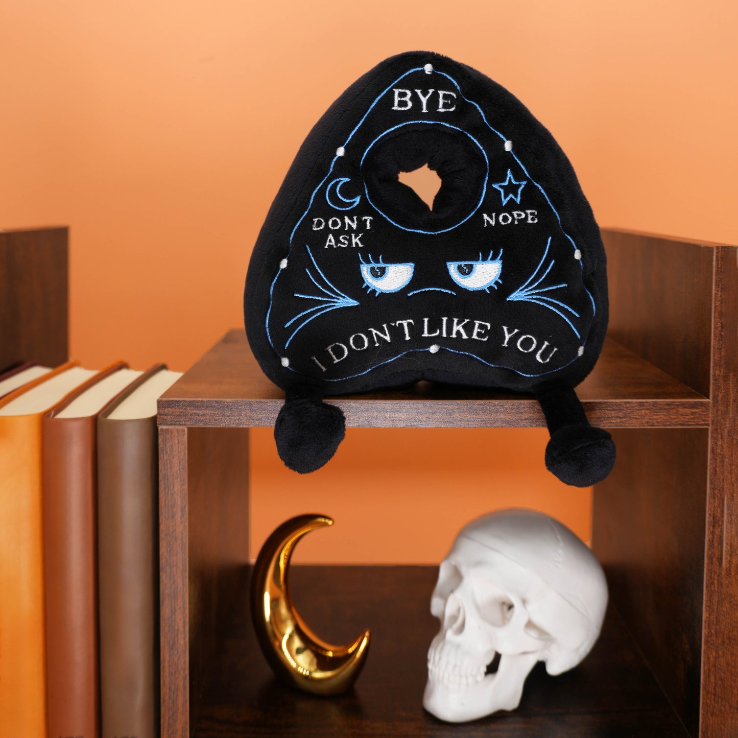 Black planchette plush pillow with text on a wooden shelf against an orange wall.