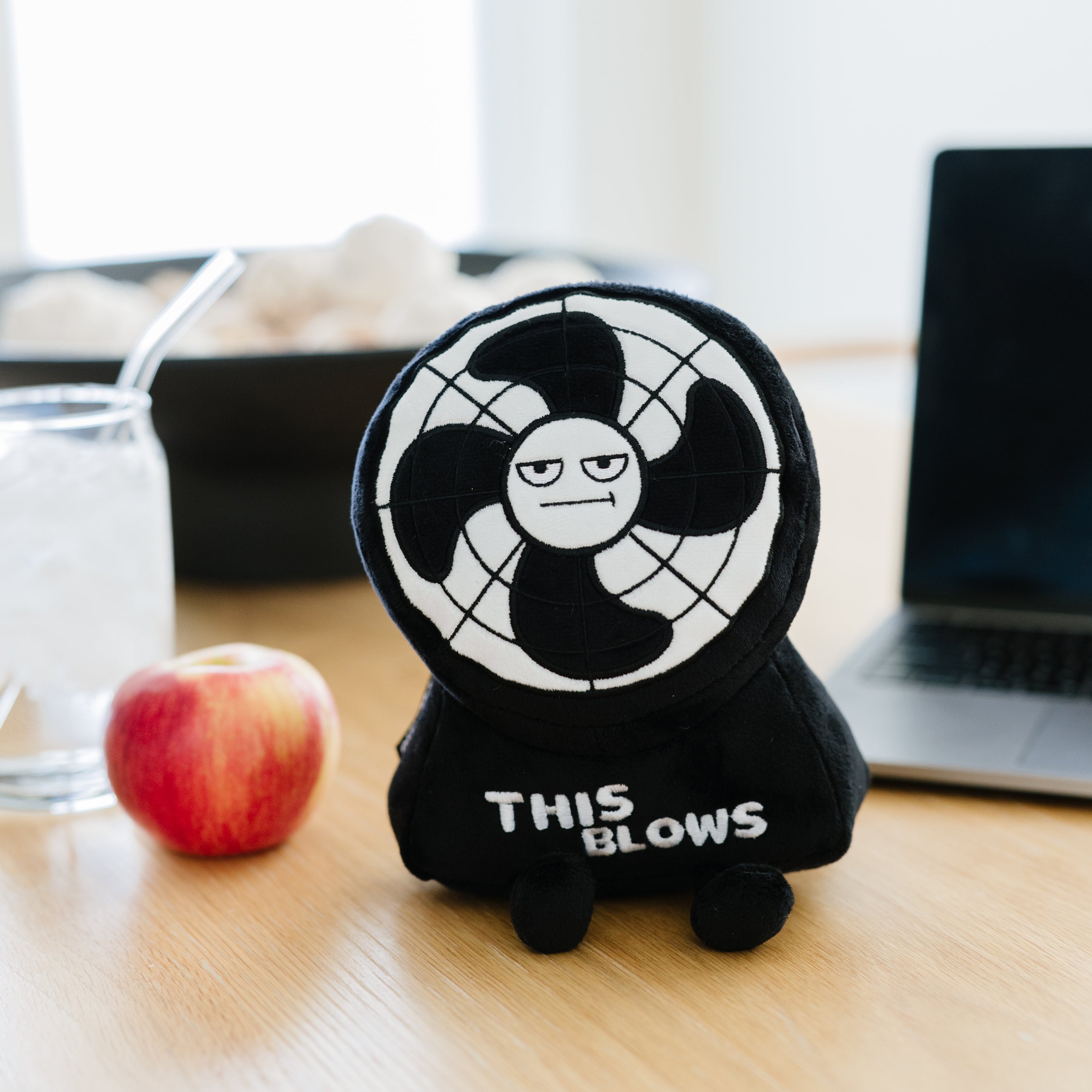 A plush in the shape of a fan with a grumpy expression and the text 'This Blows' on its base placed on a kitchen table.