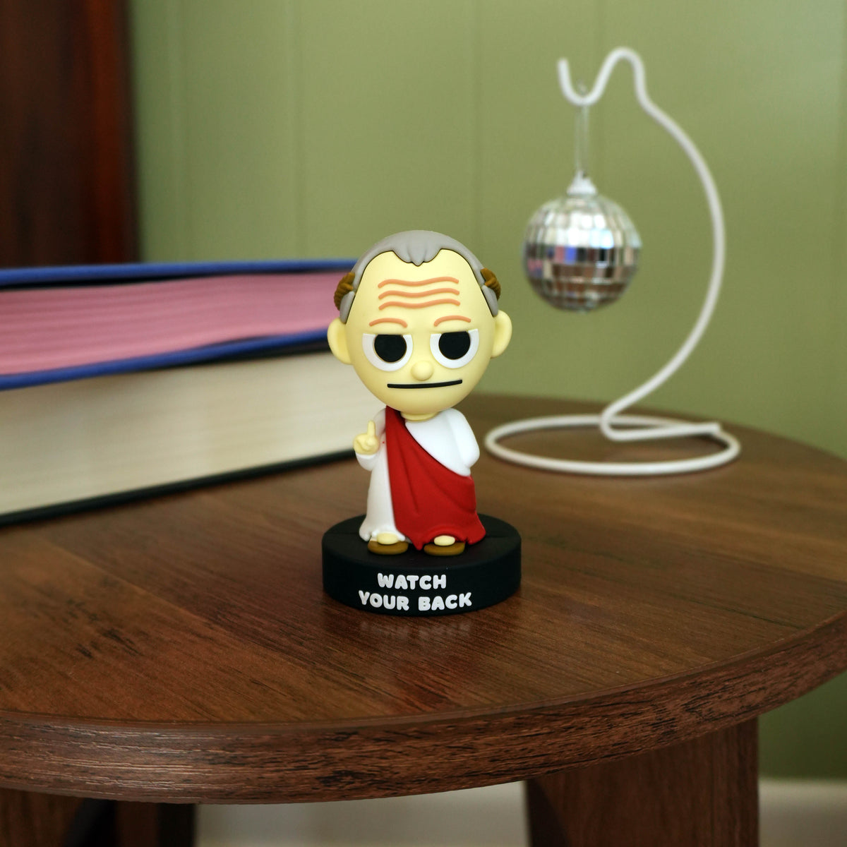 A small bobblehead figurine of Ceaser stands on a black base that reads 'WATCH YOUR BACK' on a wooden table. A book, green wall, and silver disco ball stand in the background.