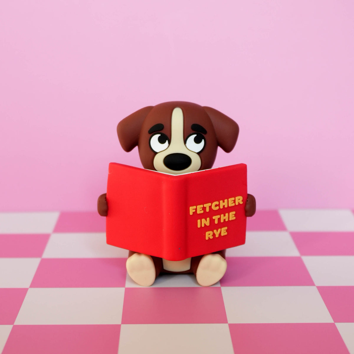 A small dog figurine with brown and cream fur holds a red book with yellow text reading 'FETCHER IN THE RYE' against a pink background with a pink-and-white checkered floor.