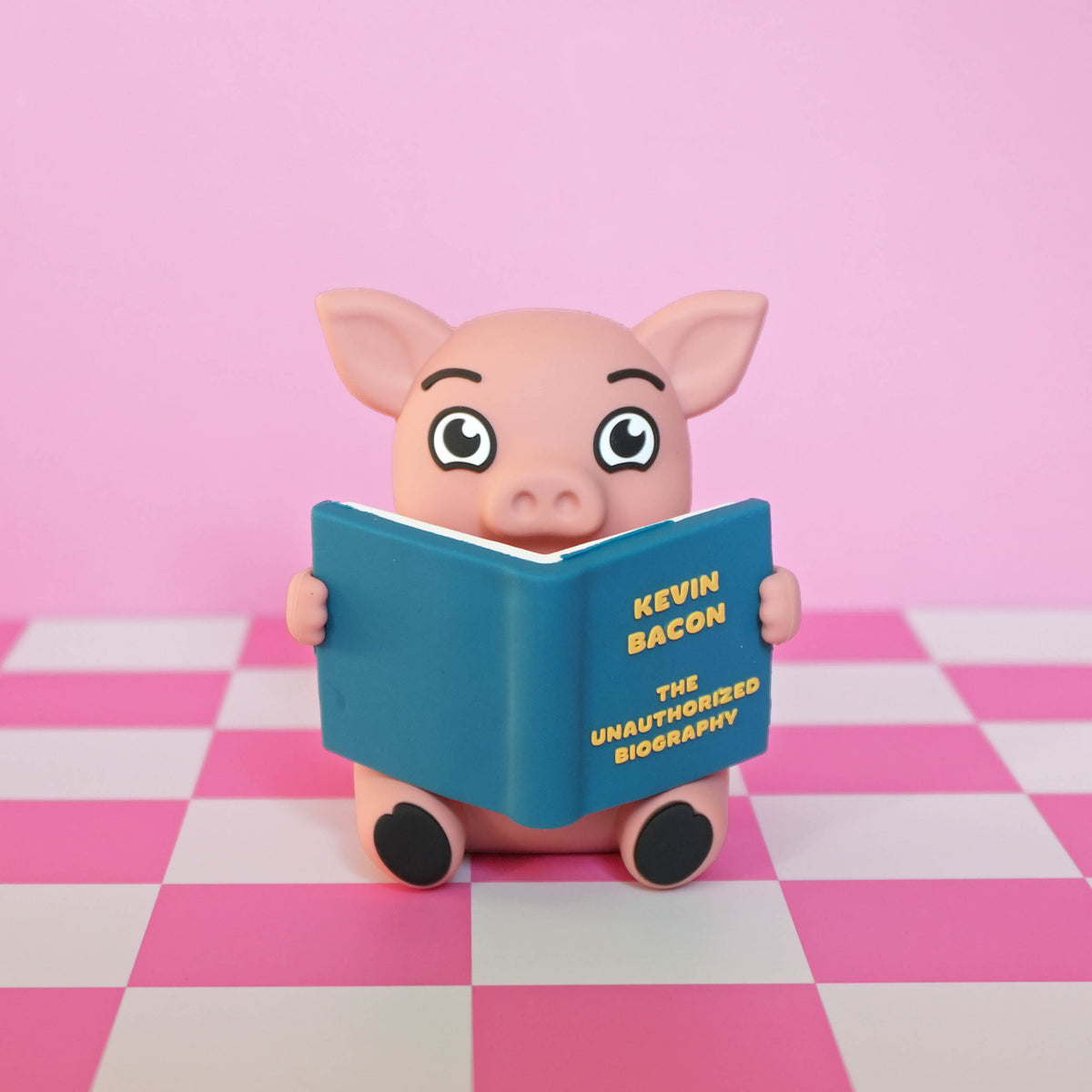 A pink pig figurine holding a blue book titled 'Kevin Bacon: The Unauthorized Biography' against a pink and white checkered floor and pink background.