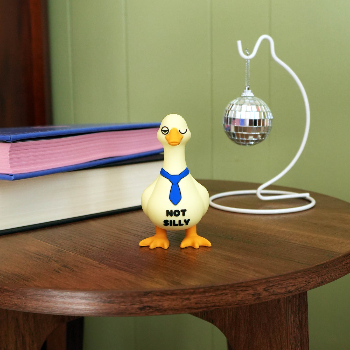 A small goose-shaped novelty figure standing on a wooden table, with the text 'NOT SILLY' printed on its front. The goose is cream-colored with orange feet and beak, and wears a blue tie.