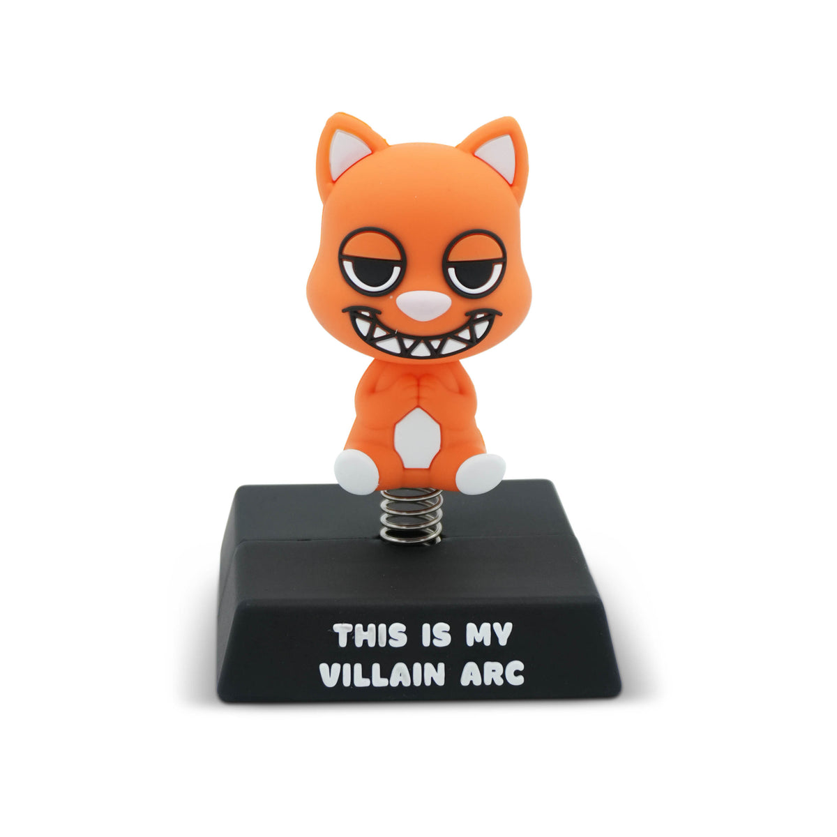 An orange cartoon cat figurine with a grinning toothy expression, mounted on a black rectangular base that reads 'THIS IS MY VILLAIN ARC' in white text. The cat sits on a metal spring, photographed on a clean white background.