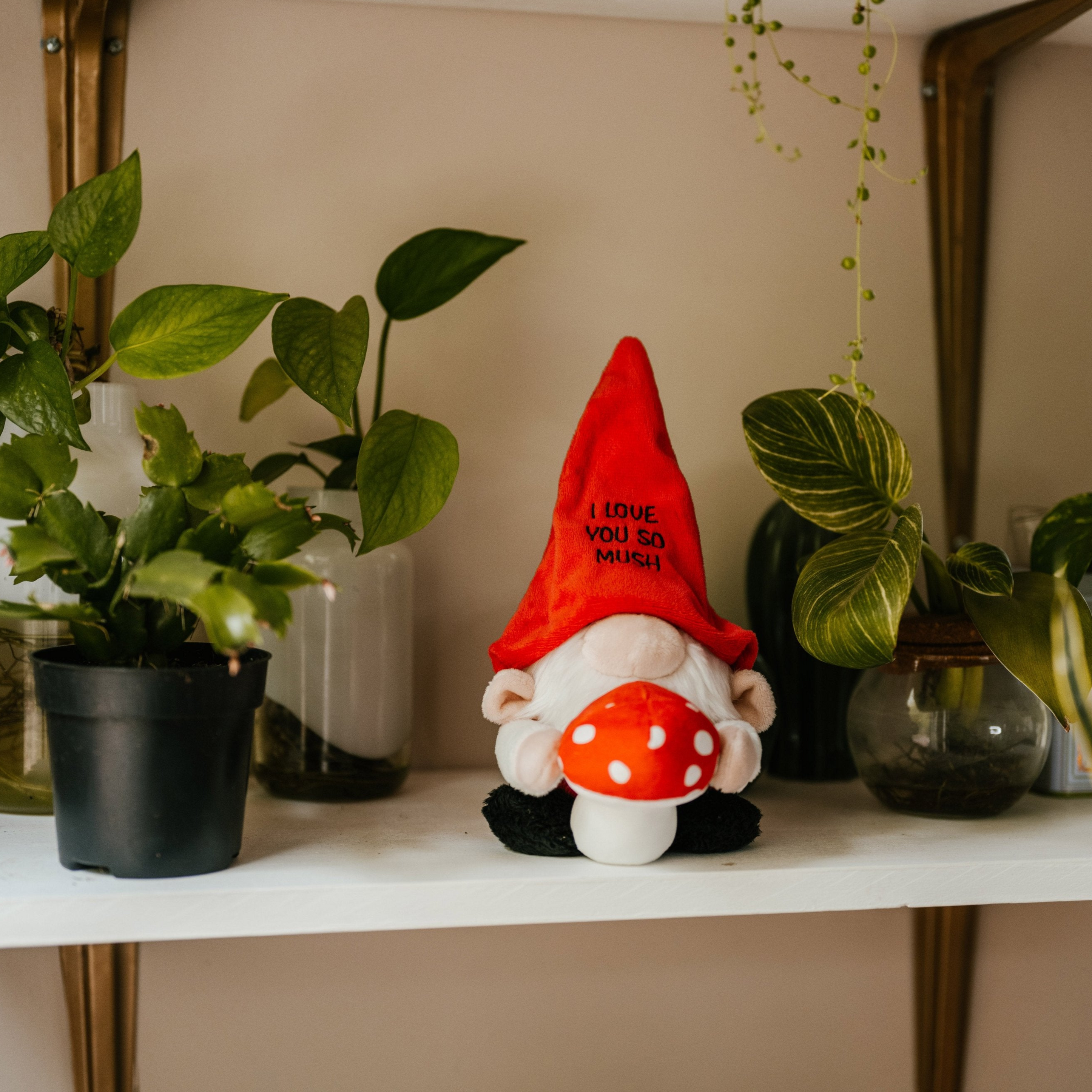 A plush gnome with a red hat with 'I love you so much' text written on it is holding a 3D mushroom. It's placed on a shelf with plants in the background.