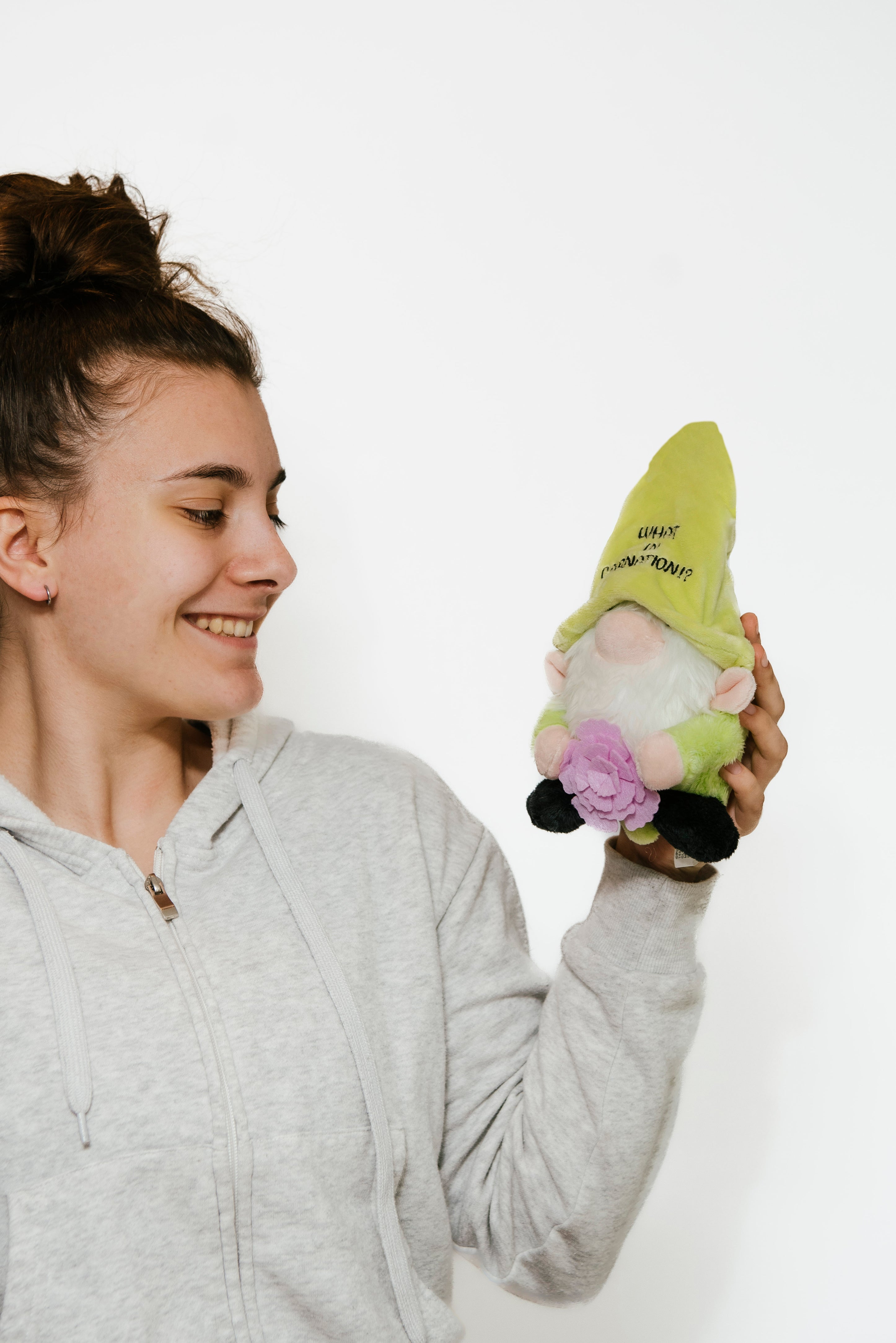 A model holding a plush gnome with a green hat with the text 'WHAT IN CARNATION' written on it, holding a purple flower.