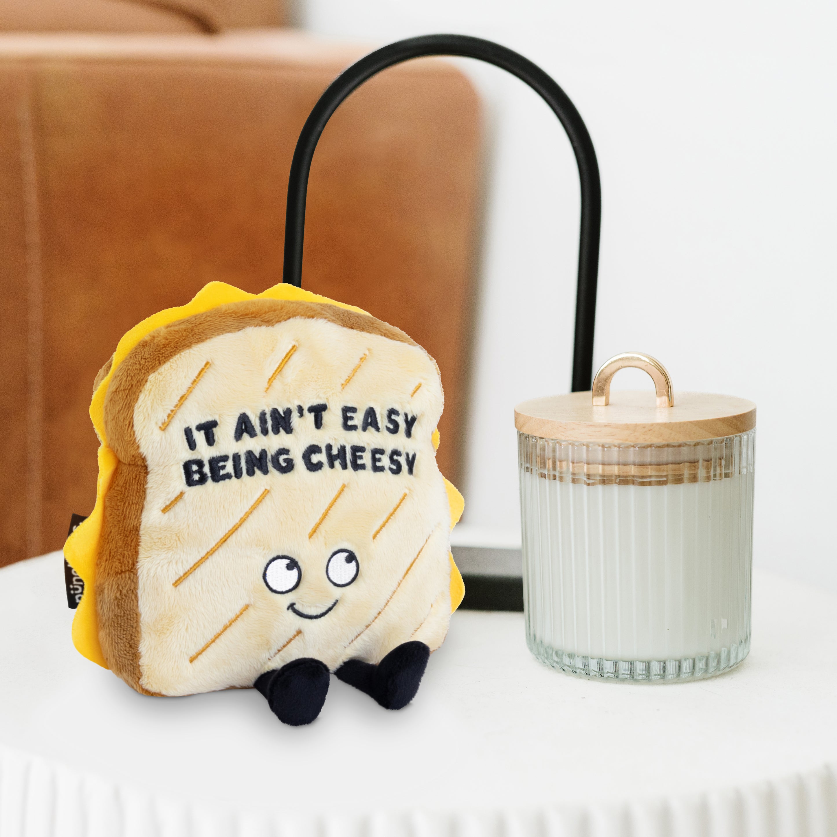 A plush toy designed to look like a grilled cheese sandwich with the phrase 'IT AIN'T EASY BEING CHEESY' written on it placed on a table in a living room.