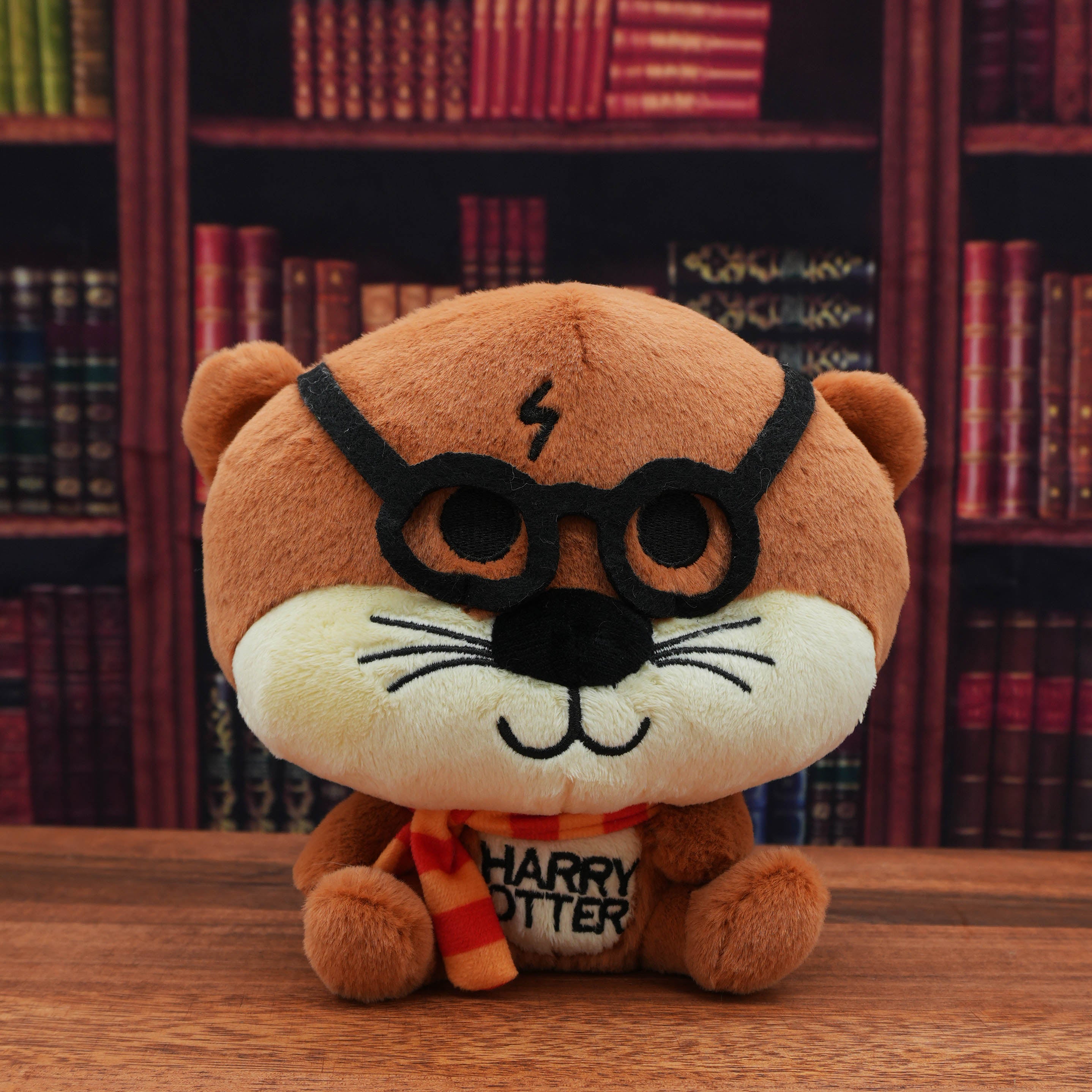 Plush toy of an otter resembling a character from 'Harry Potter' with glasses and a scarf, sitting on a wooden surface with bookshelf in the background.