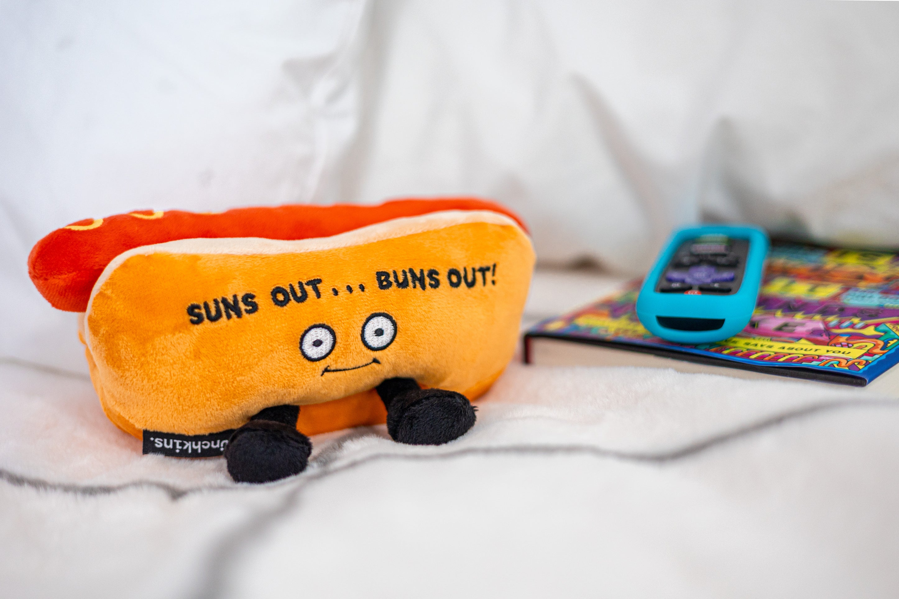 A plush shaped like a hot dog with the text 'SUNS OUT....BUNS OUT placed on a couch.