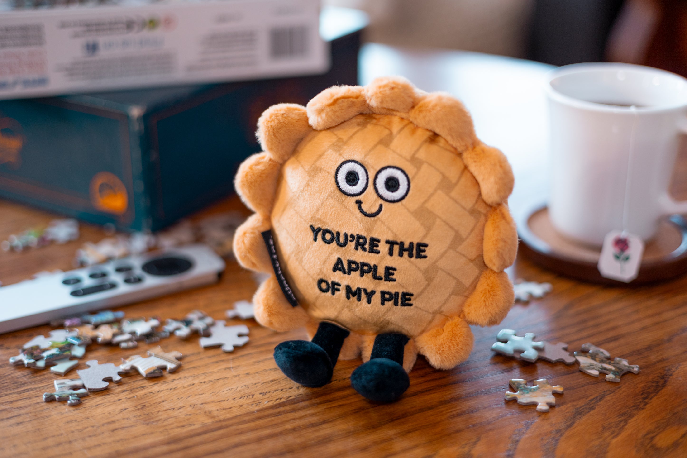 A plush designed to look like an apple pie with the phrase 'YOU'RE THE APPLE OF MY PIE' written on it. It's placed on a table near a puzzle.