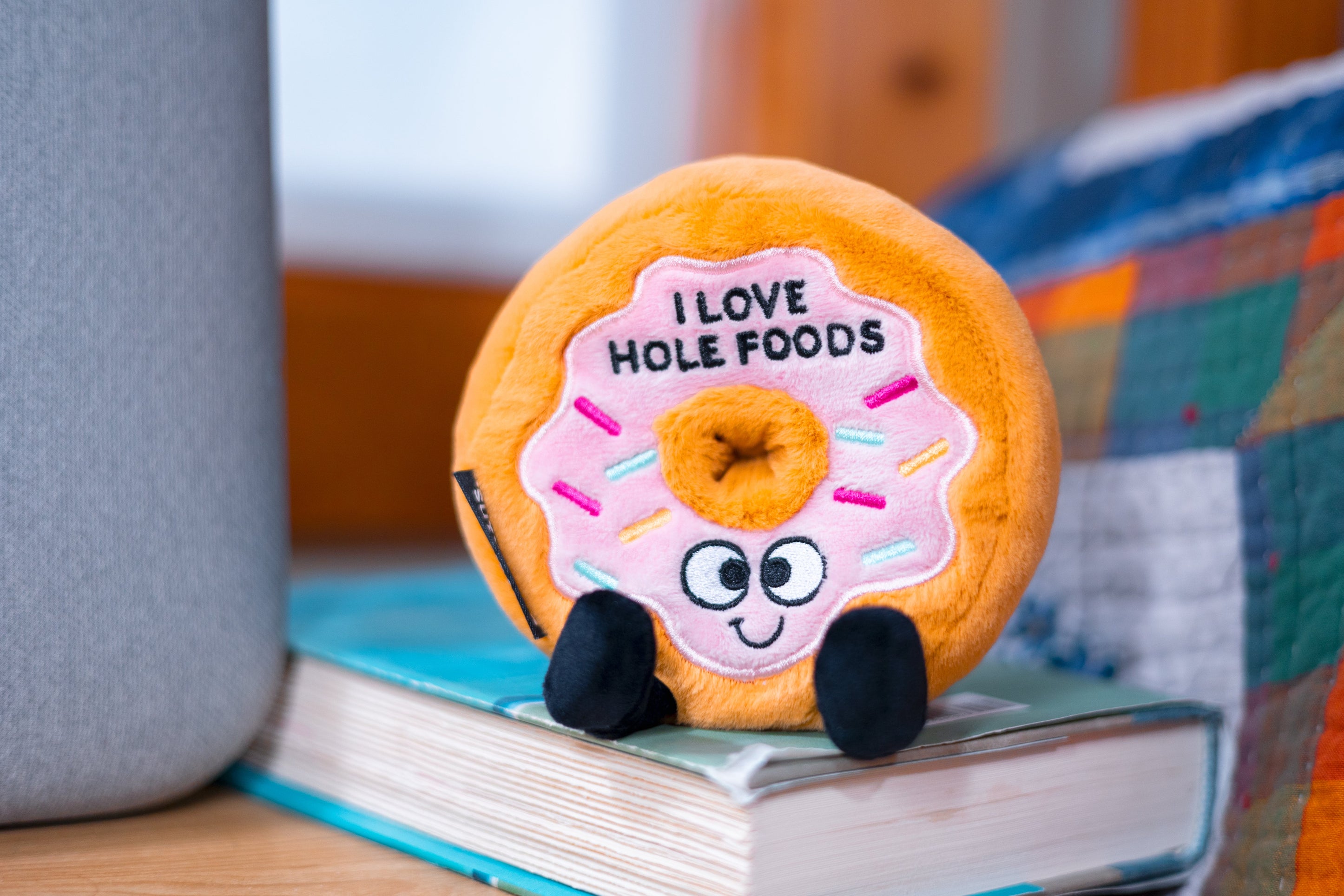 A plush designed to look like a glazed donut with sprinkles, eyes, and feet, with the text 'I LOVE HOLE FOODS' placed on a book.