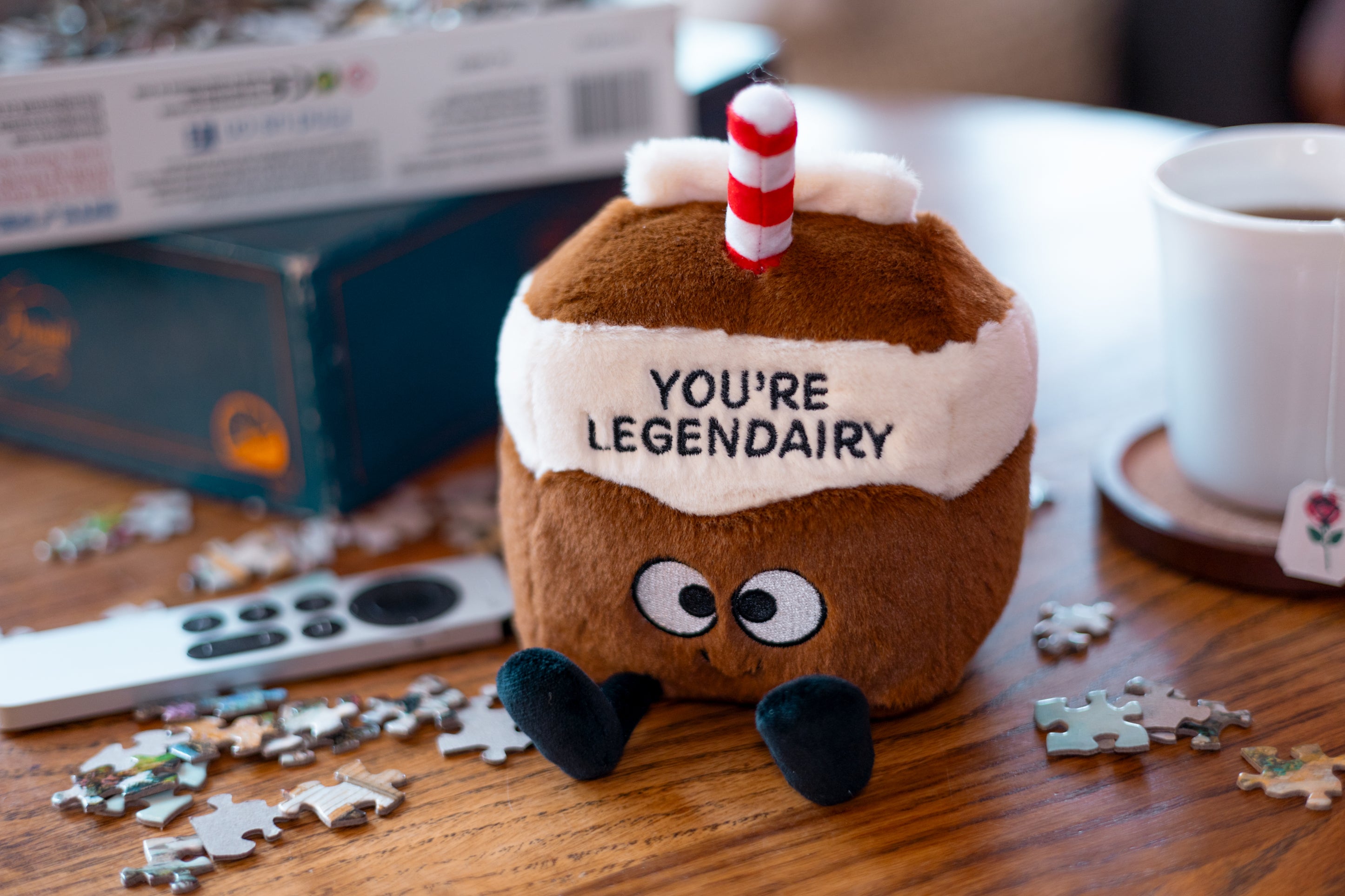 A plushie designed to look like a milk carton with brown and white colors, featuring eyes and a smile, with the text 'YOU'RE LEGENDARY' on the carton. It's placed on a table near a puzzle.