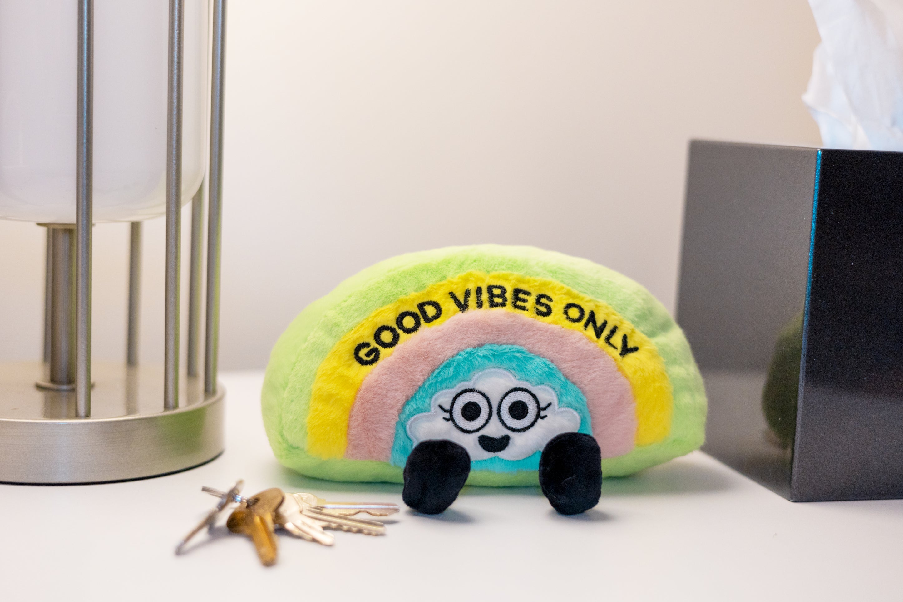A plush with a rainbow design and the text 'GOOD VIBES ONLY' on it, featuring expressive eyes and dangly legs. It's placed on a dresser.