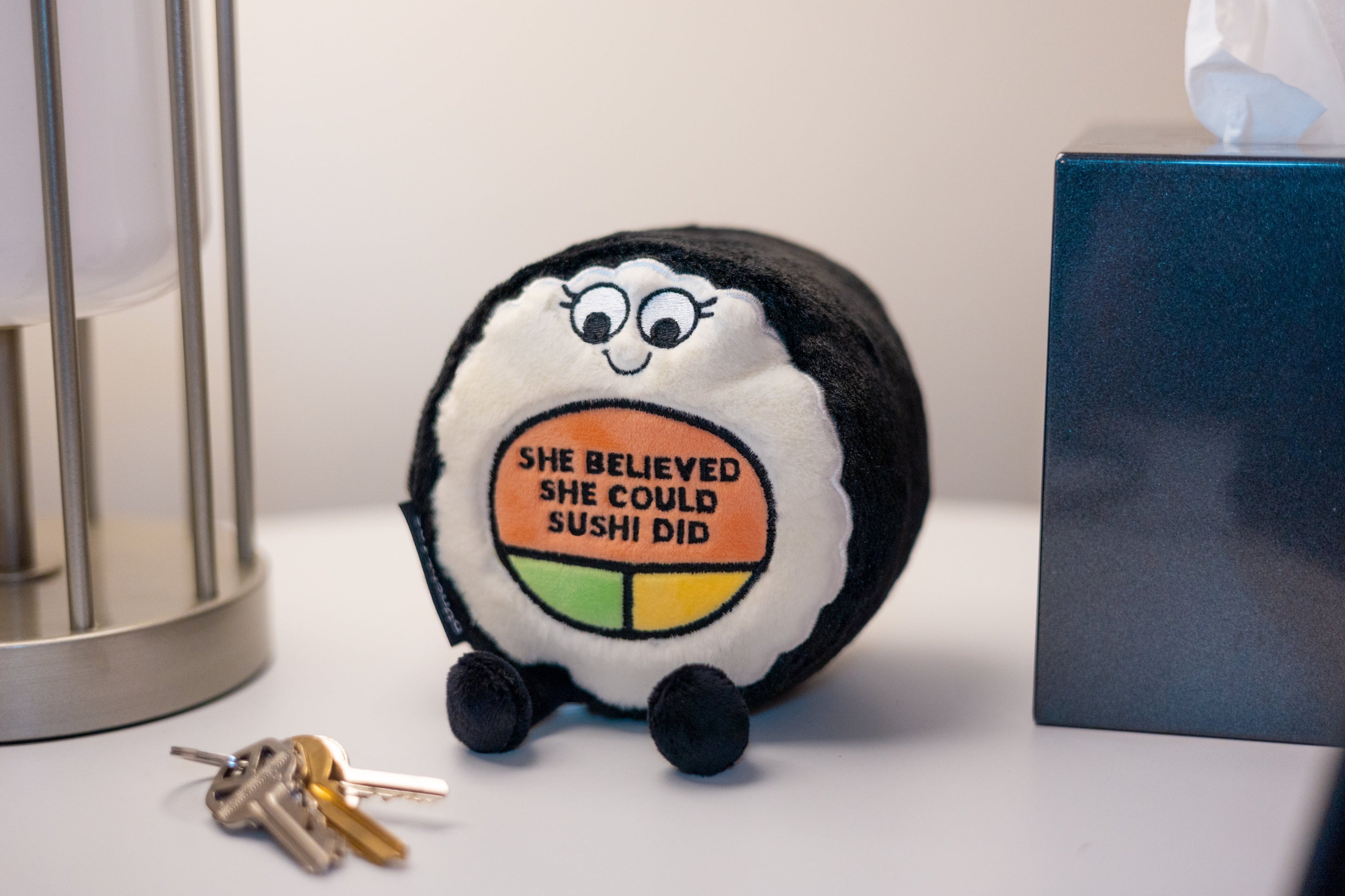 A plush designed to look like a piece of sushi with a smiling face and the phrase 'She believed she could sushi did' printed on it placed on a dresser.