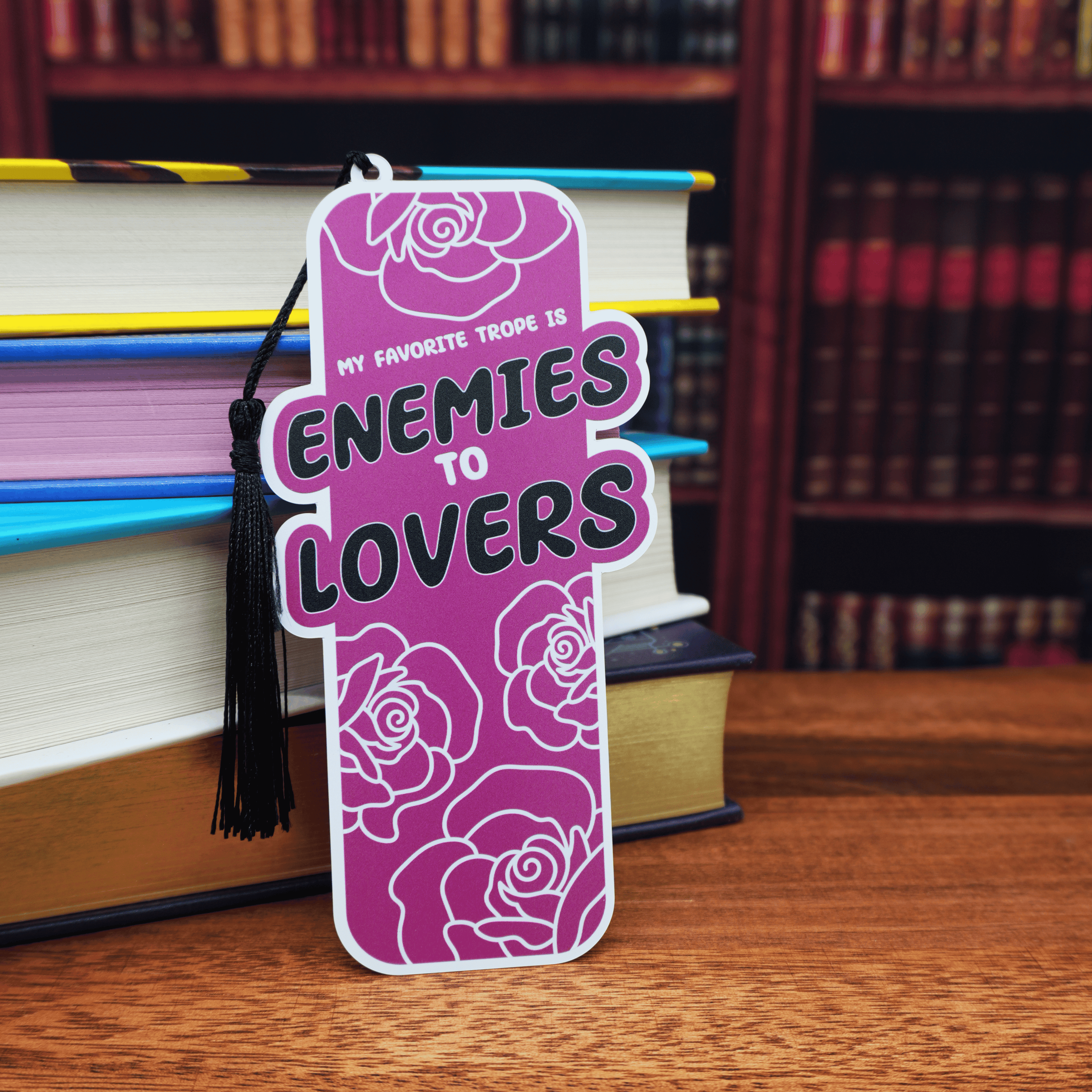 Purple bookmark with 'Enemies to Lovers' text on a wooden surface with books in the background