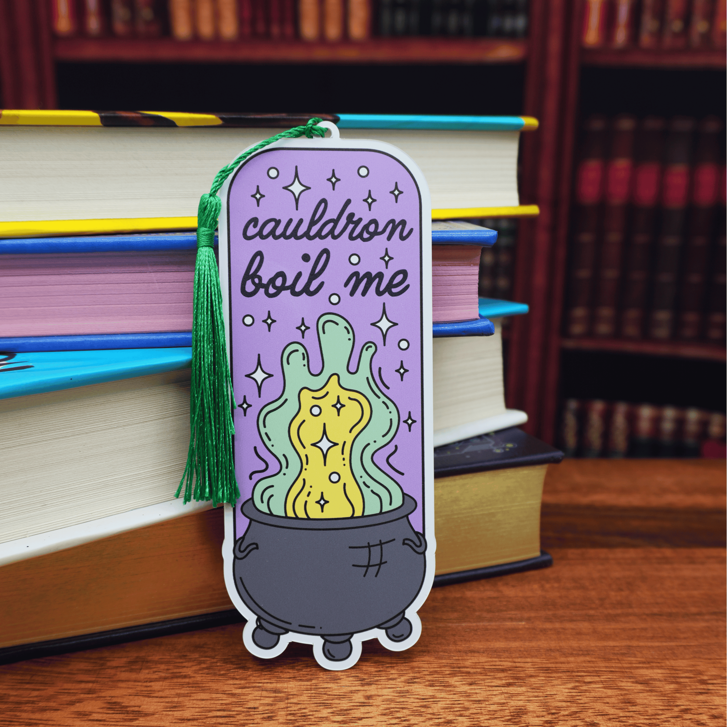 Colorful bookmark with a cauldron boil me design and text, placed on books with a bookshelf background.