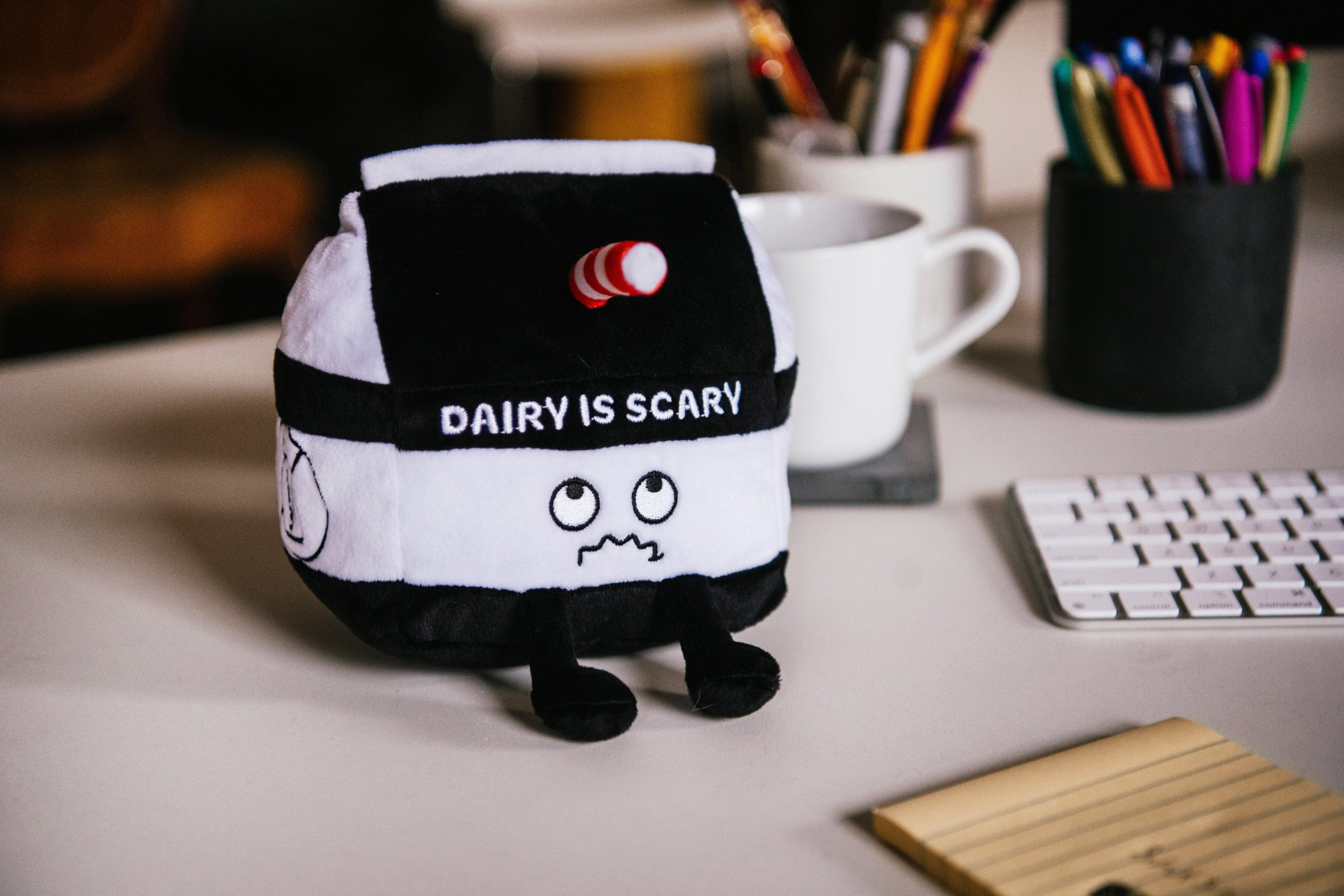 A plush designed to look like a milk carton with a distressed face and the text 'DAIRY IS SCARY' on the front placed on a desk.
