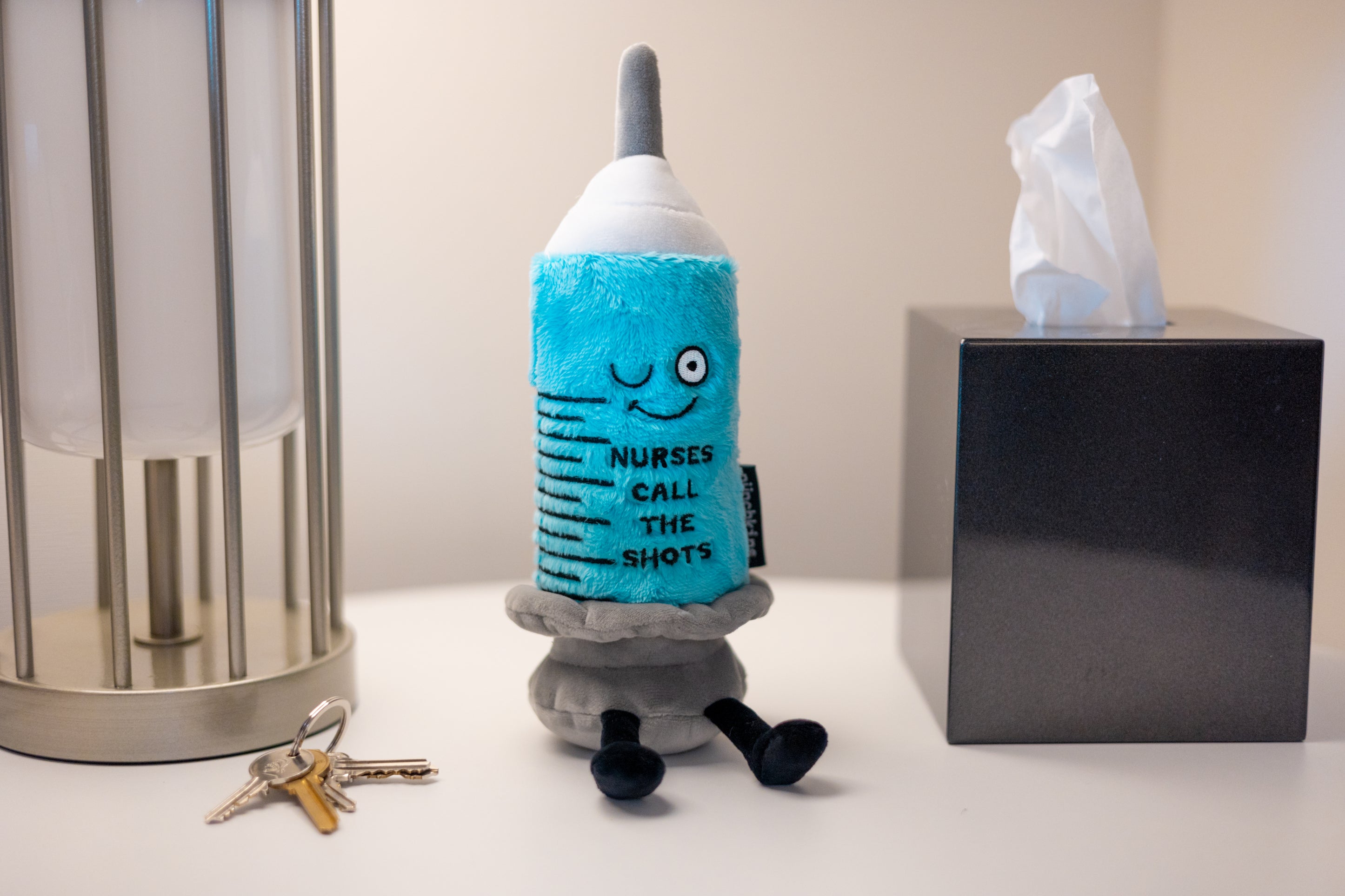 A plush designed to resemble a syringe with a smiling face and the text 'Nurses call the shot' written on it placed on a desk.