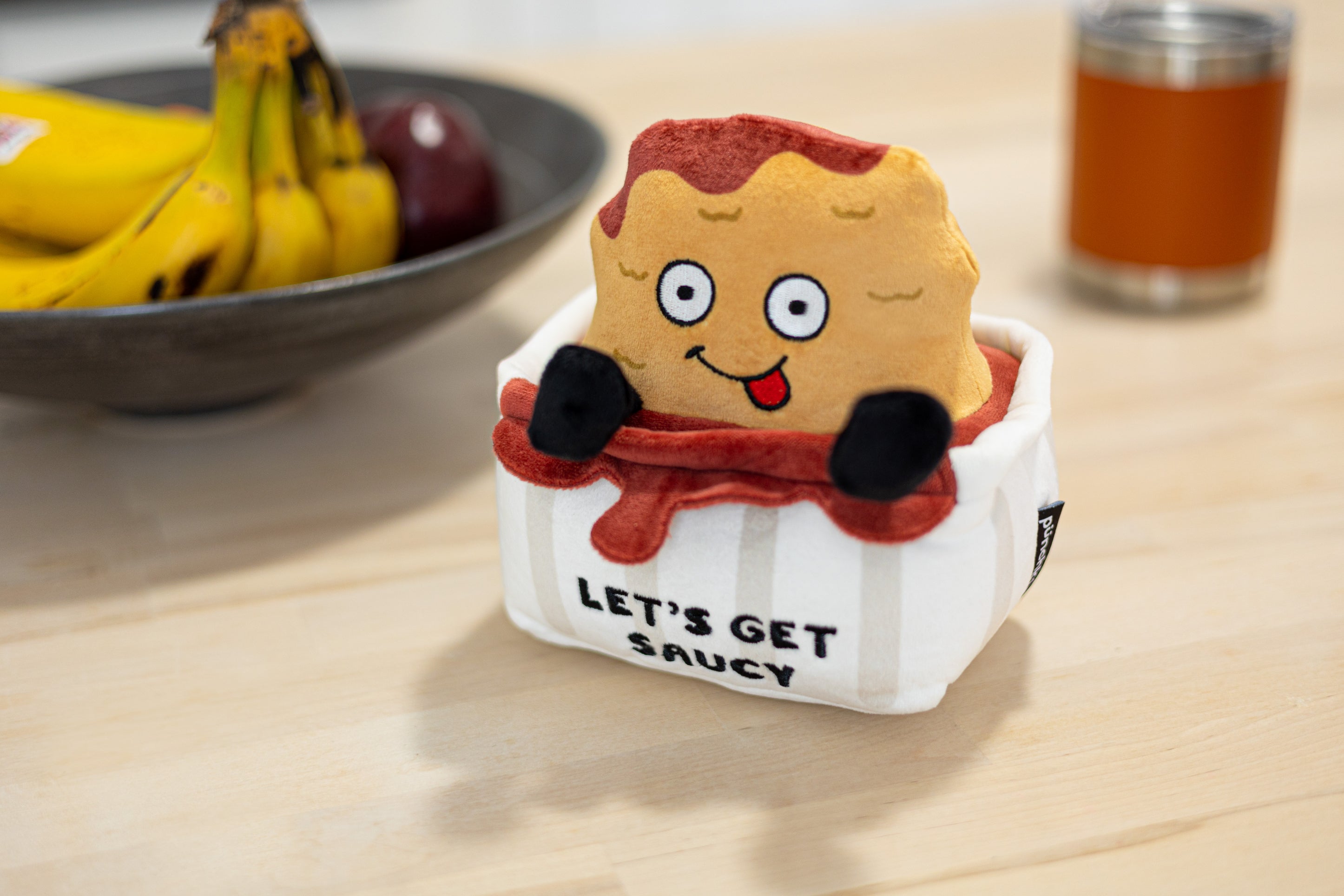 A plush designed to look like a chicken nugget sitting in a sauce container with text 'LET'S GET SAUCY' placed on a kitchen table.
