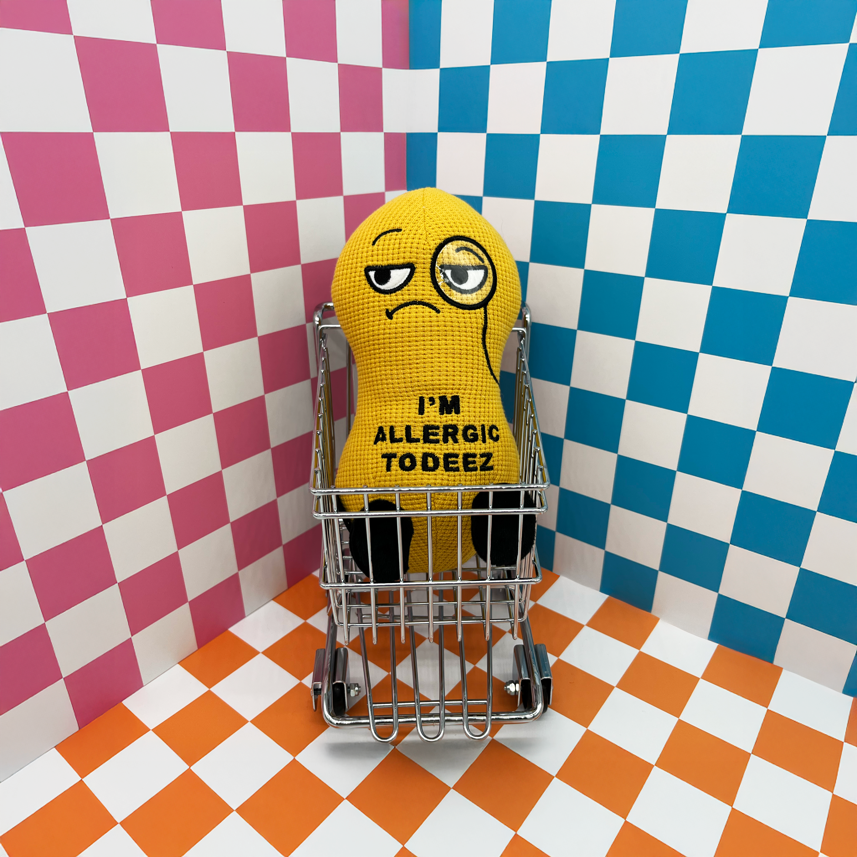 A plush shaped like a peanut with a sad facial expression and the text 'I'M ALLERGIC TO DEEZ' embroidered on its body on a checkered background in a shopping cart.