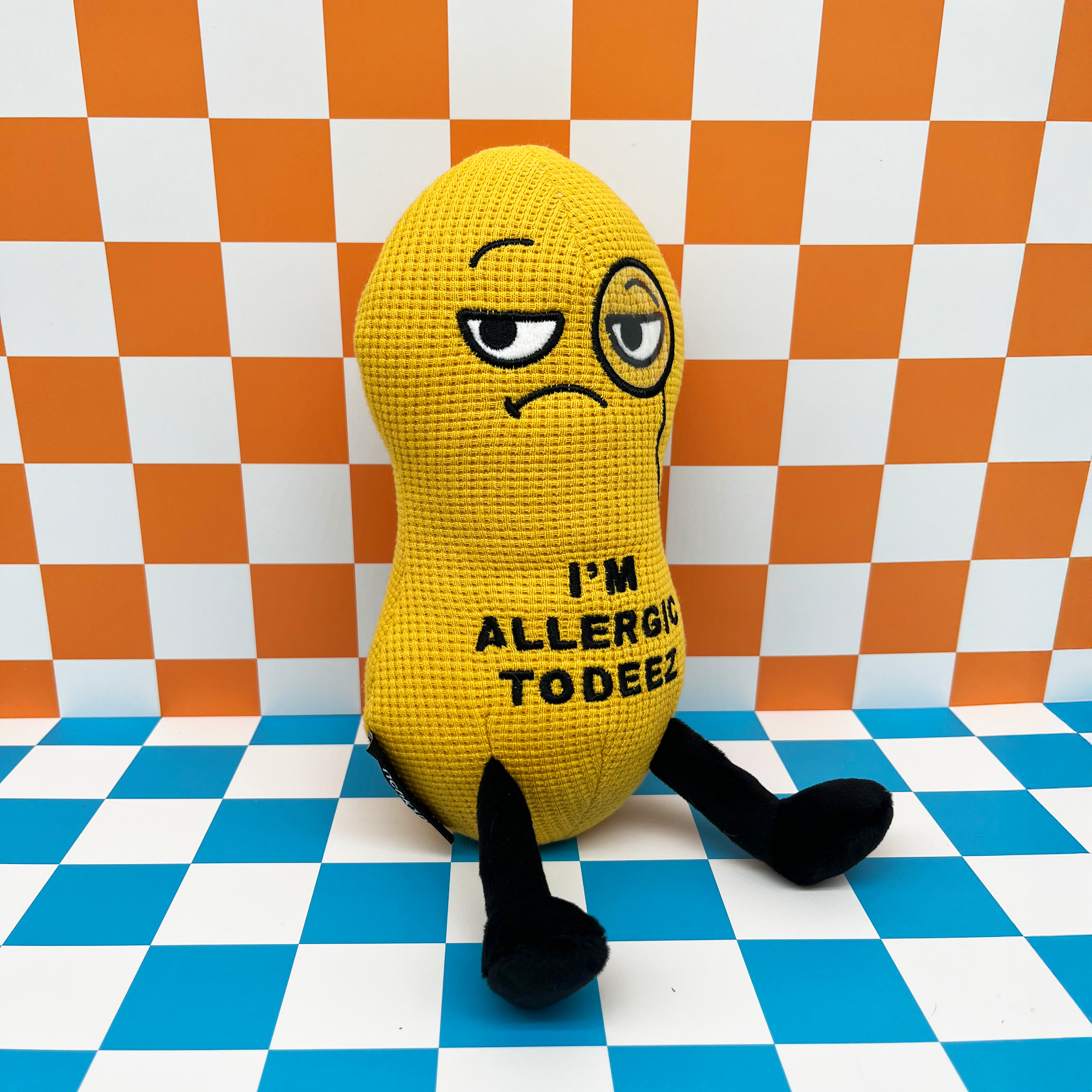 A plush shaped like a peanut with a sad facial expression and the text 'I'M ALLERGIC TO DEEZ' embroidered on its body on a checkered background.