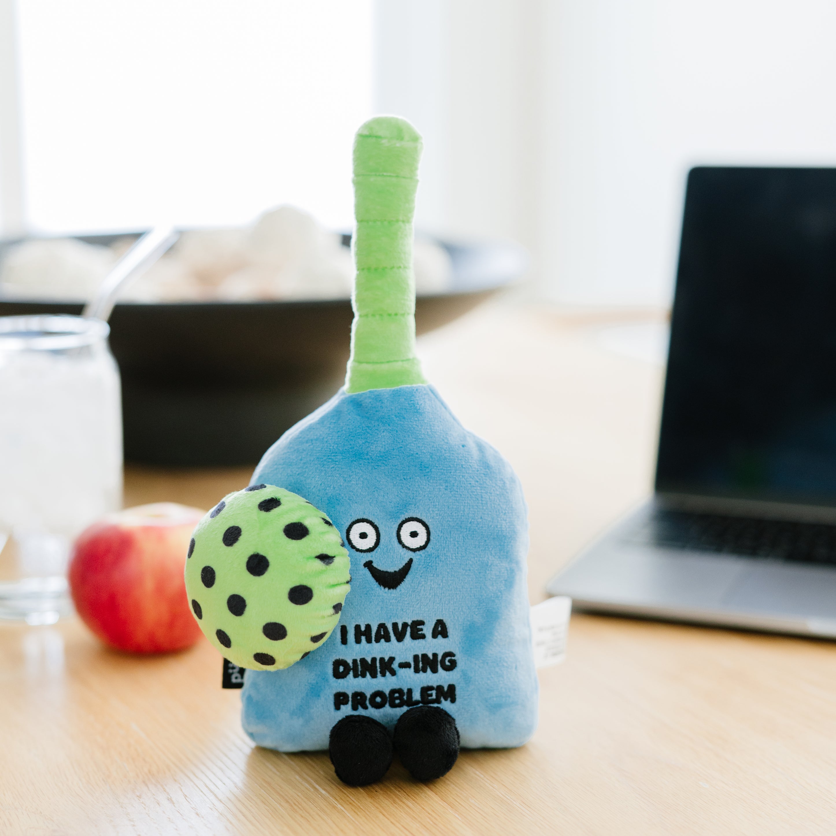 A plush pickleball paddle with 'I have a dink-ing problem' written on it placed on a kitchen table.