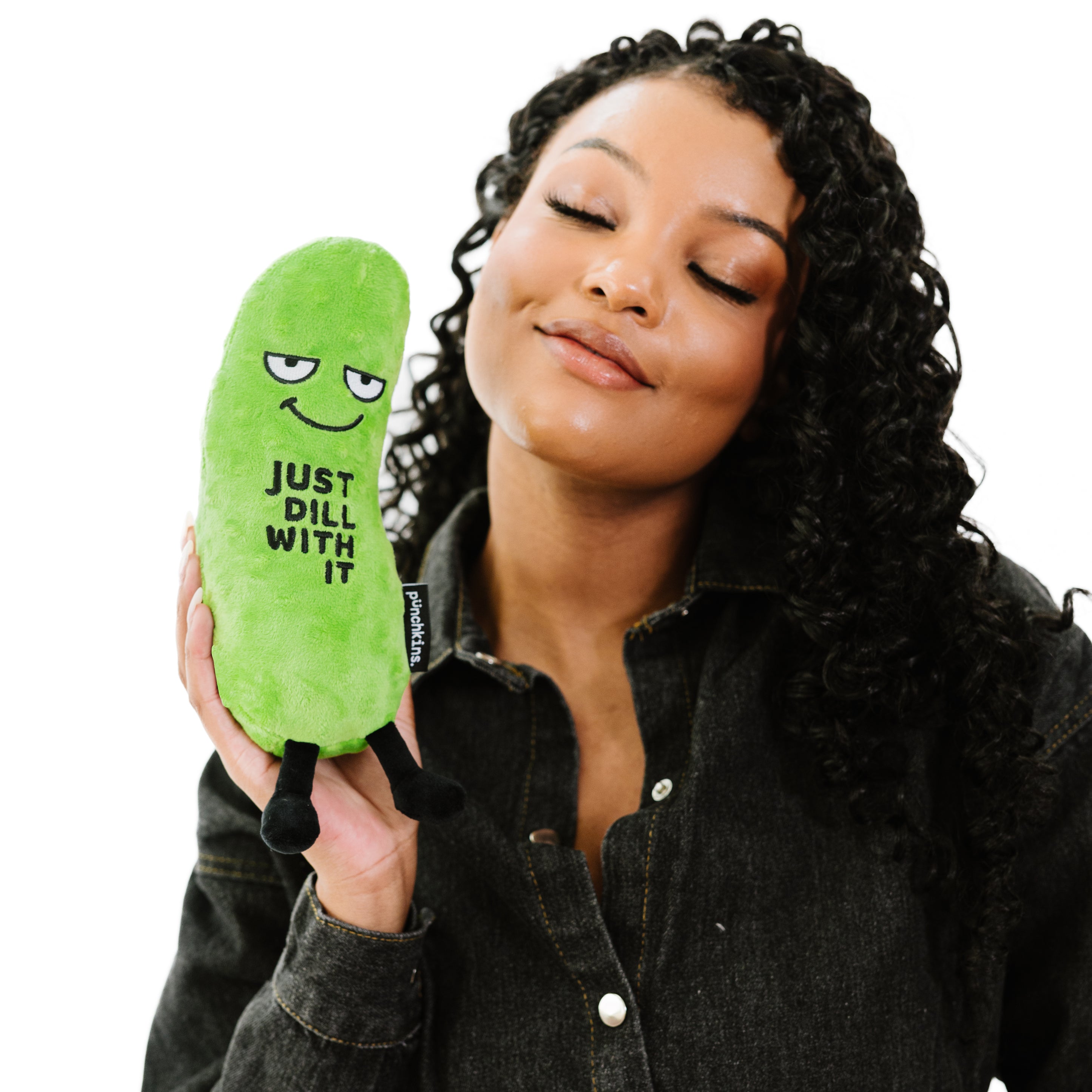 A woman holding a plush shaped like a pickle with a smiling face and the phrase 'JUST DILL WITH IT' printed on its surface.