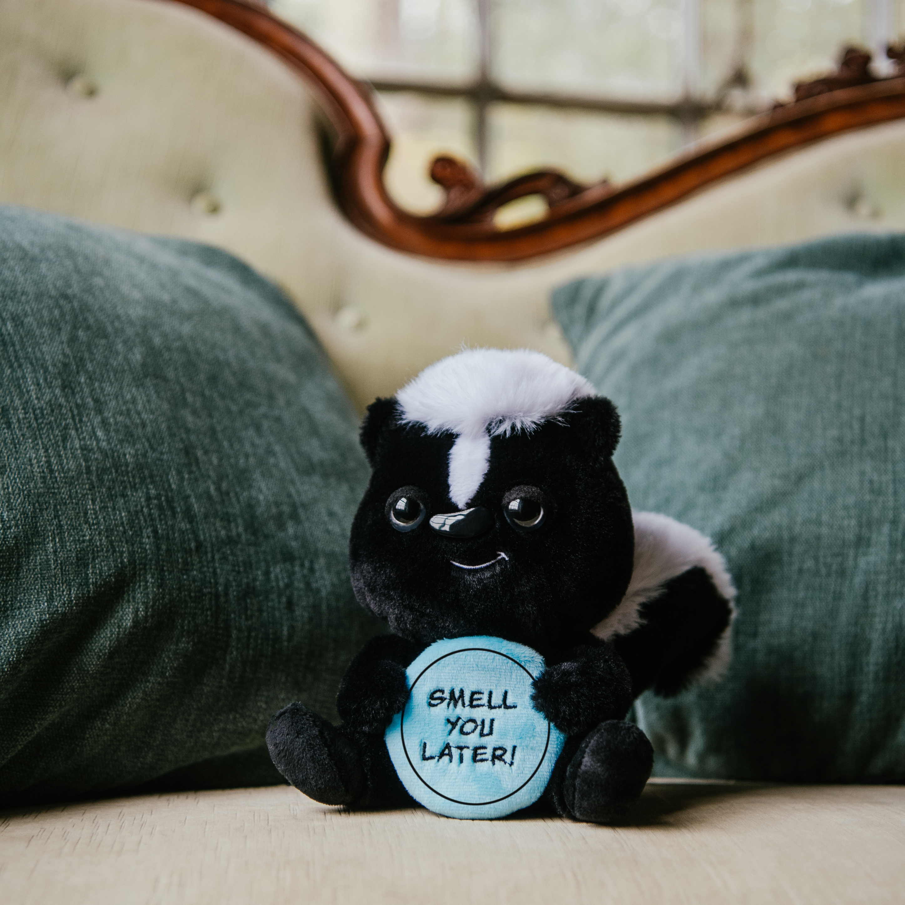 A plush resembling a skunk holding a sign that reads 'SMELL YOU LATER' placed on a couch.