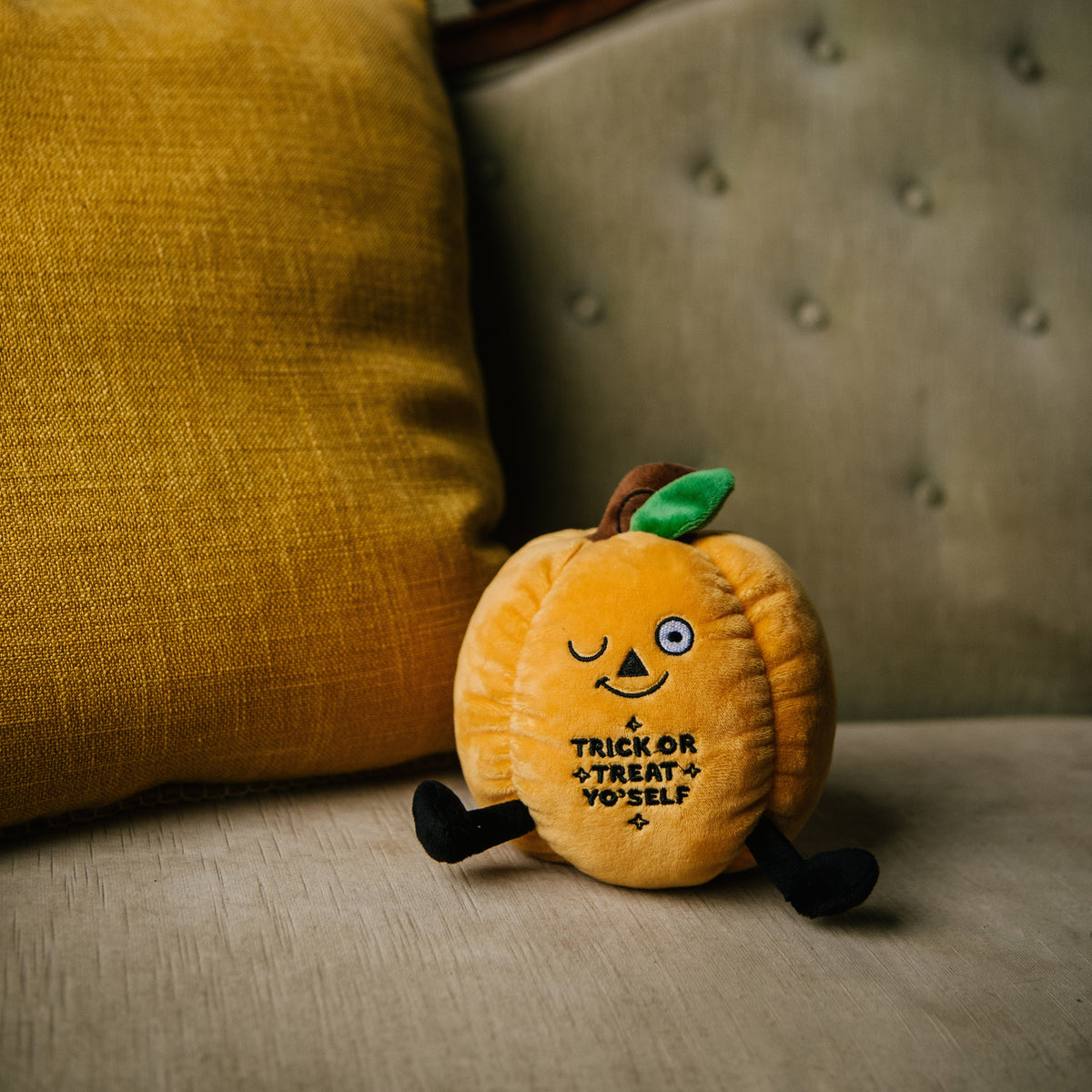 A plush designed to look like a smiling orange pumpkin with the phrase 'TRICK OR TREAT YO'SELF' written on it placed on a couch.