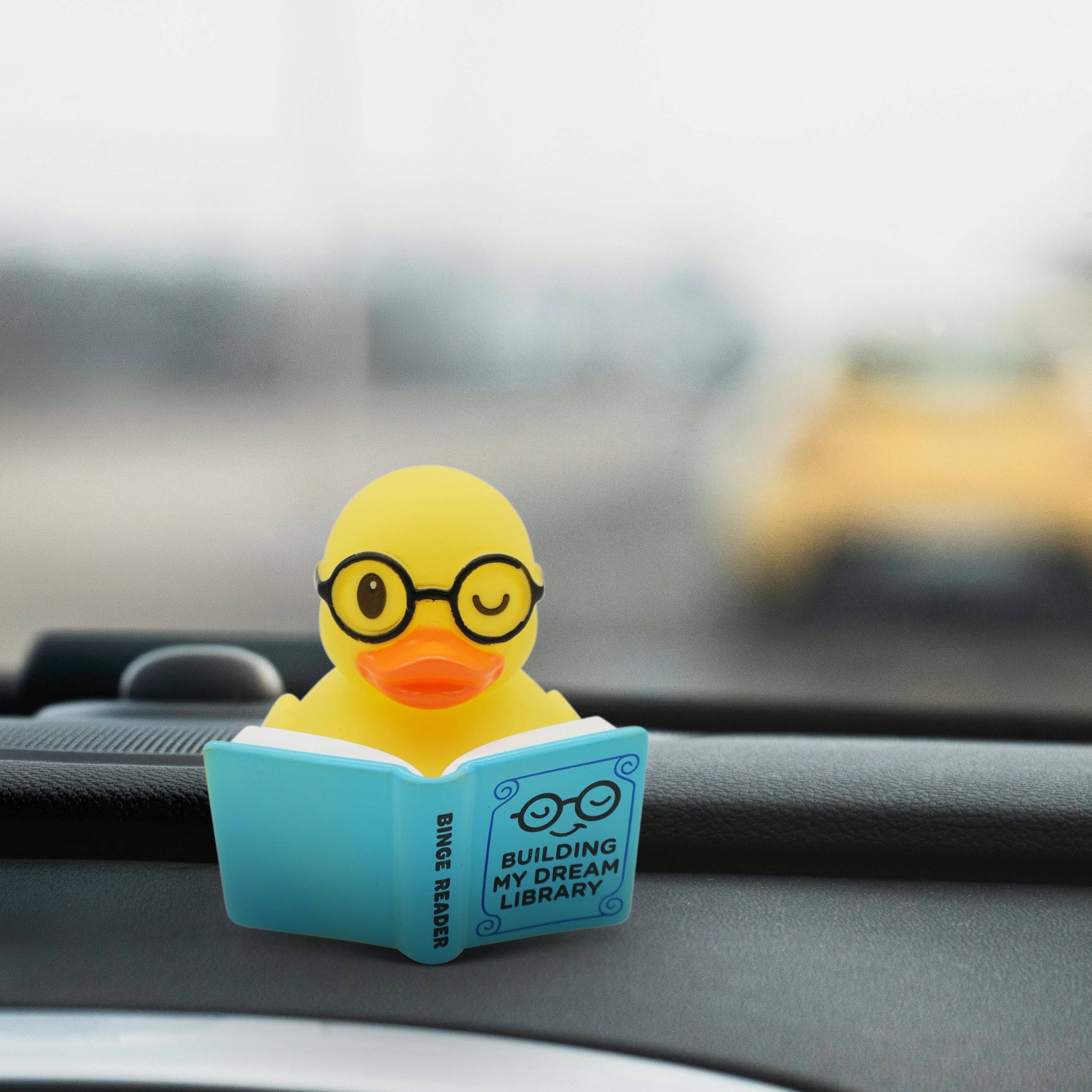 Yellow rubber duck with glasses reading a book on a car dashboard